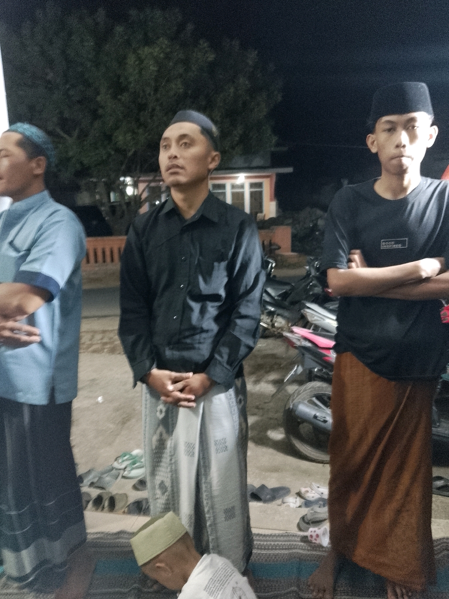 Three young males, all wearing traditional Islamic attire, stand in an outdoor, nighttime setting in Rejoyoso, Indonesia. The male on the left wears a light blue collared shirt and a blue cap, with his arms crossed. The middle male wears a black collared shirt, a patterned sarong, and his hands are clasped in front of him. The male on the right wears a black t-shirt with a small white logo and a brown sarong, and his arms are crossed. A fourth young male, wearing a light green cap, is kneeling in the foreground, his head bowed. Several pairs of footwear are scattered on the ground behind the standing males, and a motorcycle is visible in the background. The scene appears to be a communal gathering, possibly a religious event, taking place on a dirt or gravel surface.