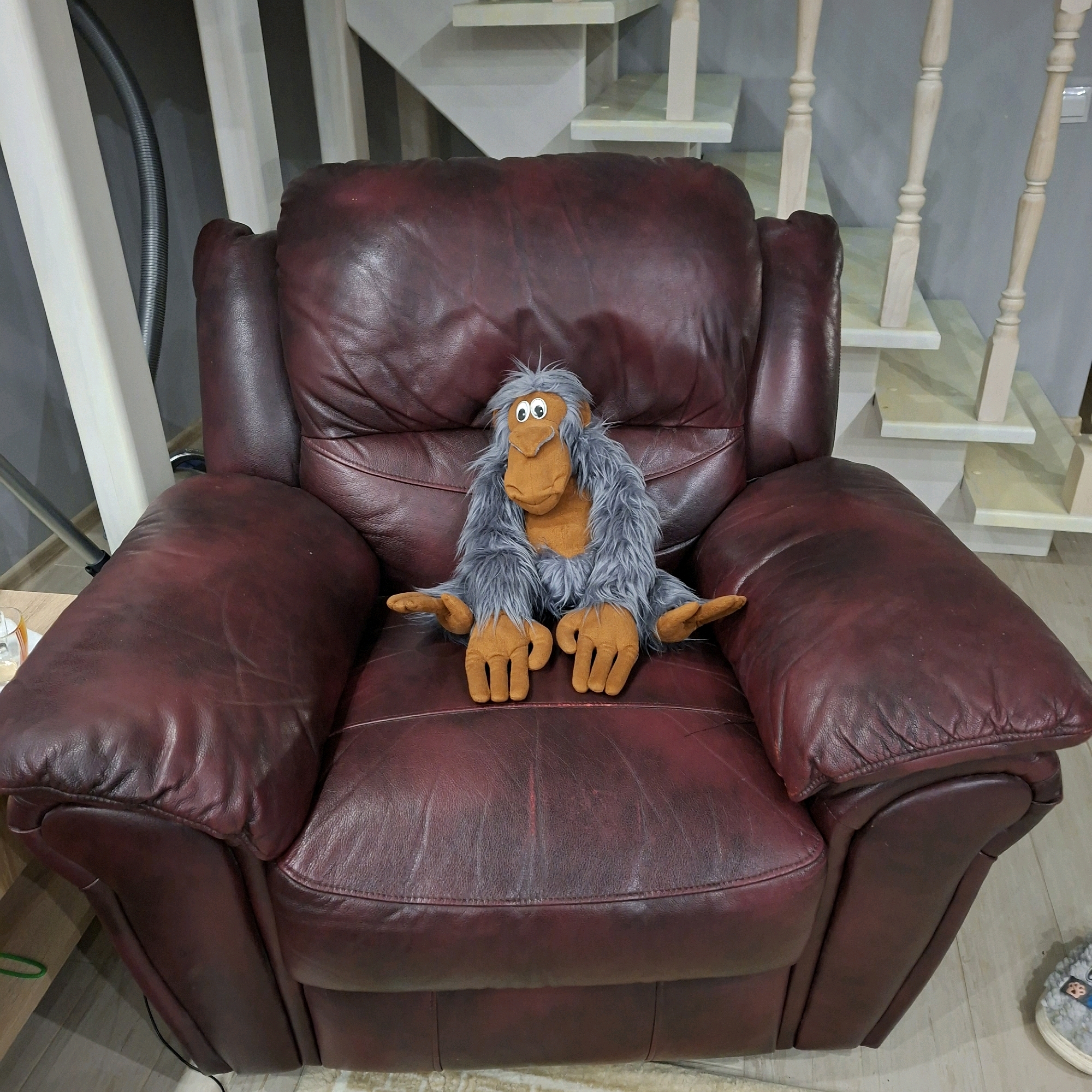 A plush toy ape with grey shaggy fur and orange hands and face sits on a dark red leather recliner. The recliner is positioned in front of a wooden staircase with white banisters and light-colored steps. The floor is a light-colored wood laminate. In the background, a grey wall is visible. To the left of the recliner, a black flexible hose is attached to something out of frame. The scene appears to be indoors, likely in a residential setting. The location is not specified in the image or available context.