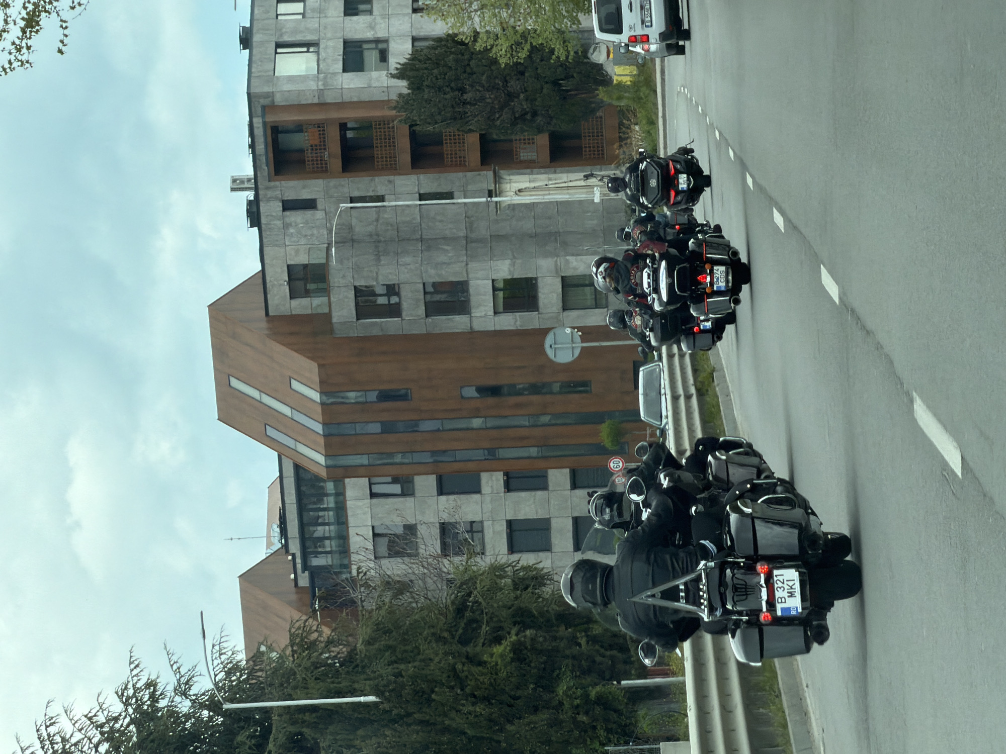 A street scene is captured, showcasing a column of motorcycles traveling on a paved road lined with white lane markings. The motorcycles are dark-colored, with riders wearing helmets and protective gear. The license plates on one of the motorcycles is “B 321 MK1.” The road is adjacent to a modern architectural structure, possibly an apartment or office building, featuring a facade of concrete and wood panels. The building has multiple windows and balconies. The environment includes trees and other greenery, and the sky is overcast with clouds. A white vehicle is visible on the road ahead of the motorcycles. The location is in Varna, Bulgaria, based on the motorcycle license plate which has a "B" prefix and the "RO" country code, which denotes Romania.