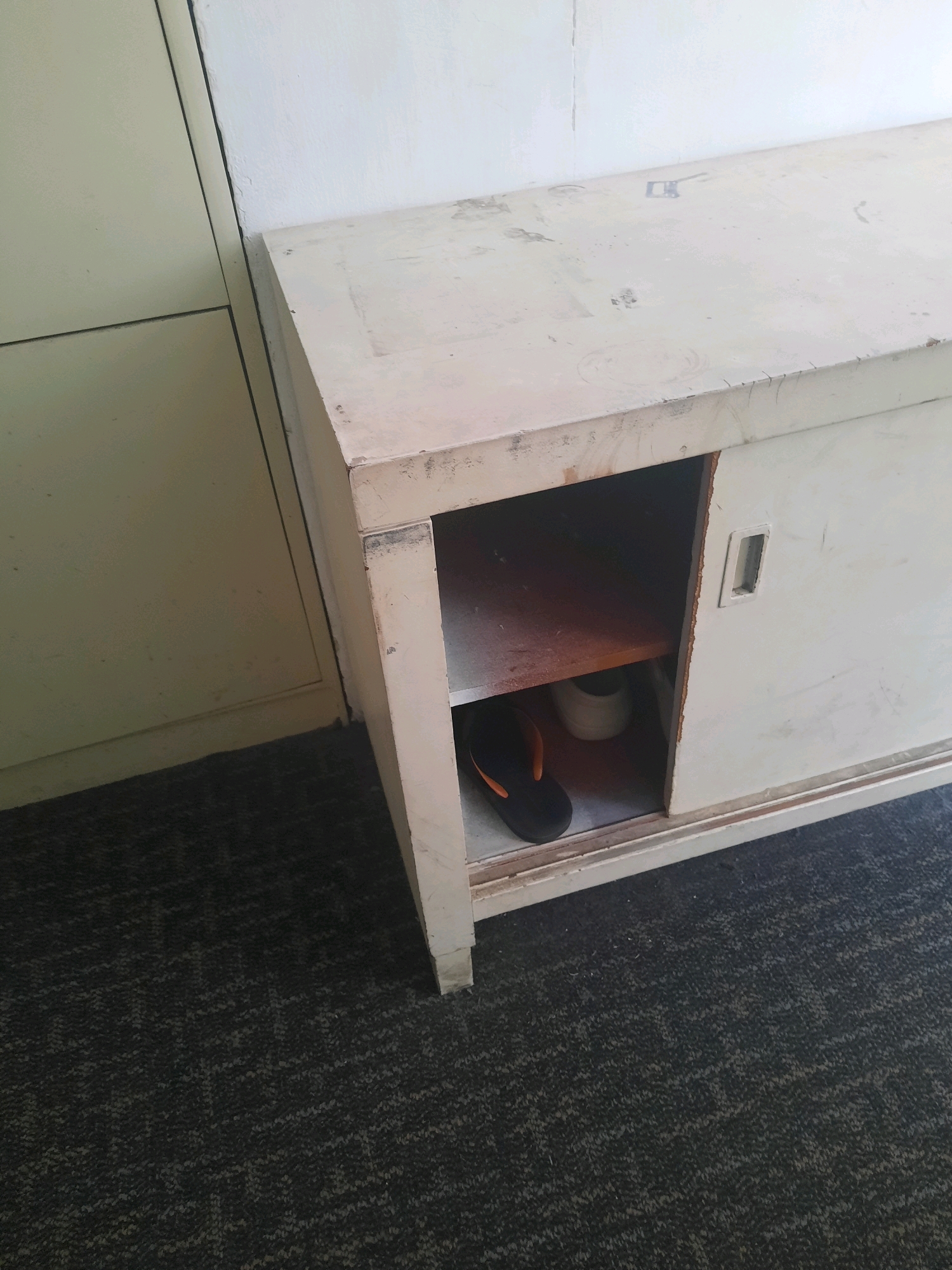 In Padang, Indonesia, a worn, off-white metal cabinet with sliding doors sits on a patterned carpet. The cabinet's top surface is marked with scuffs and faded square shapes. One of the cabinet's open compartments reveals a single black and orange flip-flop resting on a shelf, next to a white cylindrical object. The cabinet is positioned next to a larger, cream-colored vertical surface, possibly another cabinet or a wall, creating a junction where shadows are cast. The overall scene suggests an indoor, utilitarian space with signs of age and use.