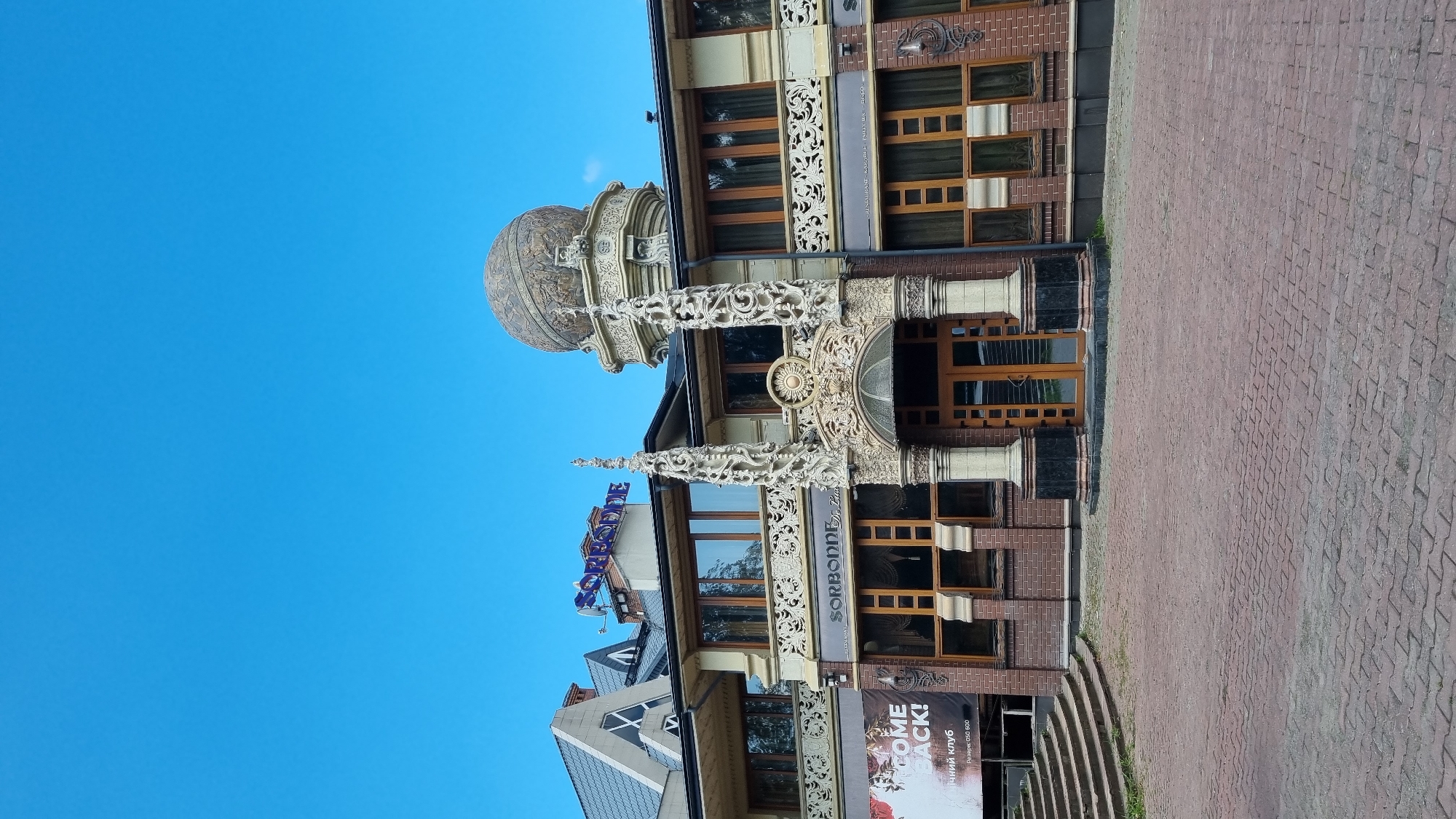 The image shows the exterior of the "SORBONNE" restaurant and lounge in Chernivtsi, Ukraine. The building features ornate architectural details, including carved stone columns, intricate friezes, and a large, decorative dome topped with a spherical object. The entrance is framed by two elaborate columns. A blue sign reading "SORBONNE" is visible on the rooftop. To the left of the entrance, a banner with the text "WELCOME BACK!" and Ukrainian text indicating "night club" is displayed. The building is constructed with brick and has multiple windows. The foreground is a paved courtyard made of red bricks. The sky is clear and blue.
