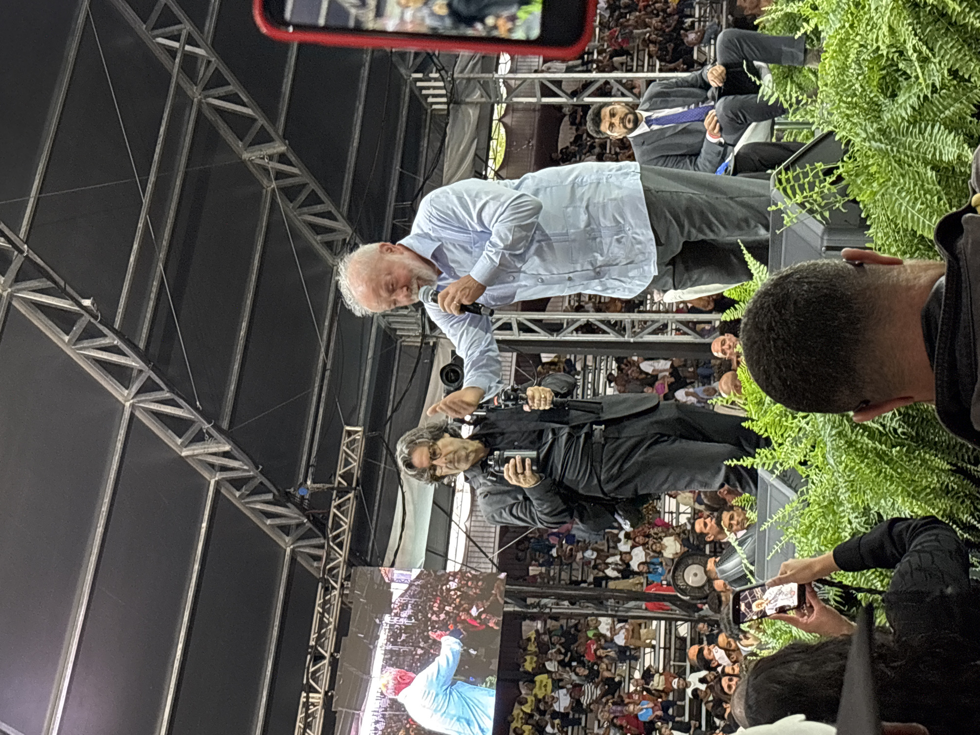 A low-angle, medium shot shows Luiz Inácio Lula da Silva speaking at an event in what appears to be São Paulo, Brazil. Lula, dressed in a light blue button-down shirt and grey pants, holds a microphone and leans slightly forward, addressing a crowd visible in the background. Beside and slightly behind him is a man in a black shirt and pants, possibly a cameraman holding equipment. A third man in a suit and tie is visible further back.
The event takes place under a metal stage structure with visible framework. A screen displays an image of a person in a blue shirt. The crowd is dense, with faces visible looking towards the stage. In the foreground, a hand holds a phone taking a picture, and a potted plant sits to the right of the frame.
