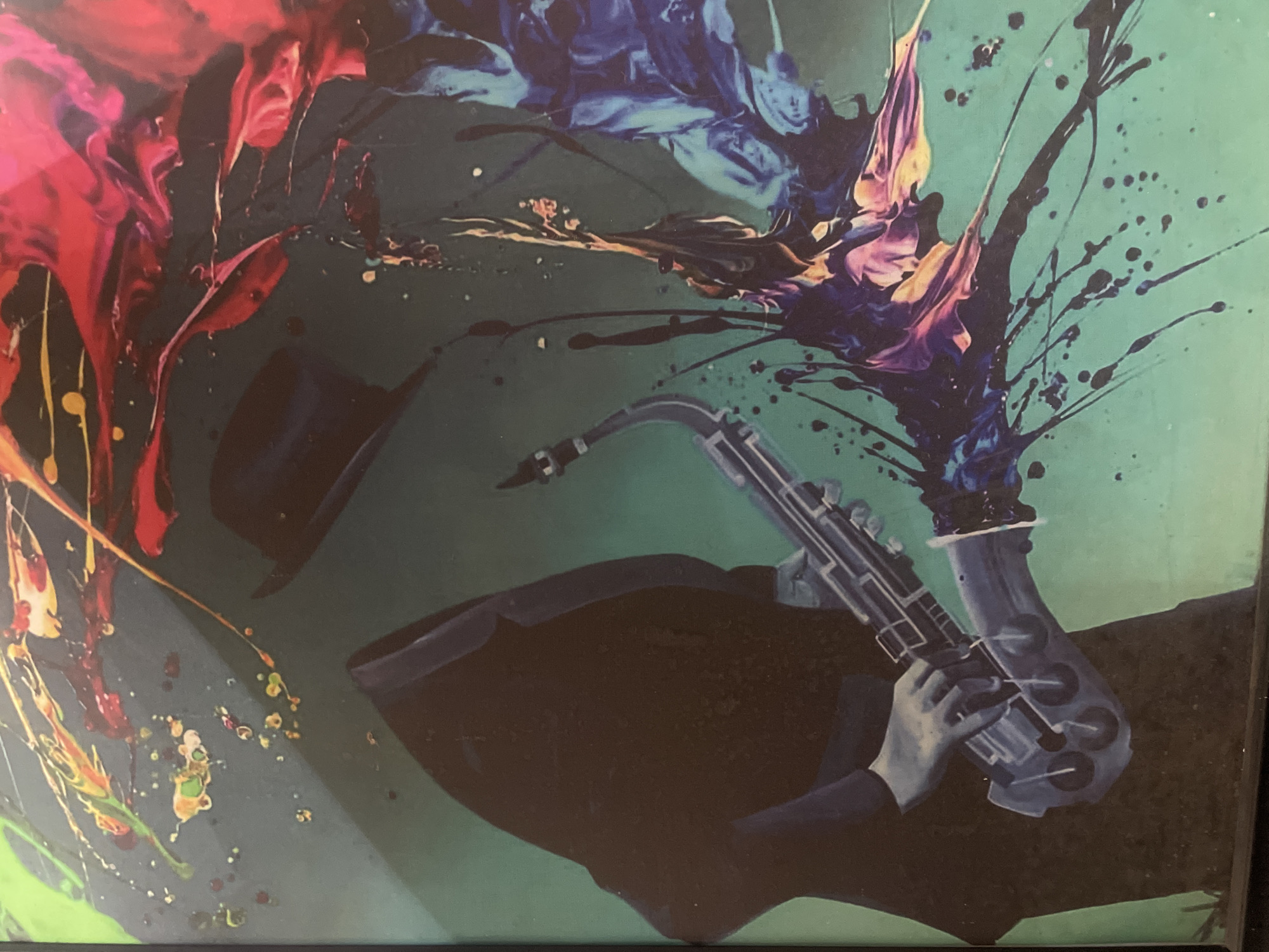 This is a vibrant and abstract artwork depicting a stylized, faceless figure playing a saxophone. The figure is largely rendered as a dark silhouette, wearing a top hat that floats above its implied head, and what appears to be a suit. Only the hands are clearly visible, gripping the saxophone, which is depicted in shades of blue-gray with noticeable keys and pads.
The background is a dynamic explosion of colors, predominantly a deep teal or dark green base. Vivid, swirling abstract forms in bright reds, magentas, blues, purples, and yellows emanate from and surround the central figure and instrument. These colorful splashes are characterized by fluid lines, drips, and splatters of paint, creating a sense of energetic movement and sound. The overall style blends figurative elements with abstract expressionism, conveying the vibrant and expressive nature of music, possibly jazz or blues. The composition suggests an outpouring of musical passion. No text is visible in the image. This artwork is observed in Abuja, Nigeria.