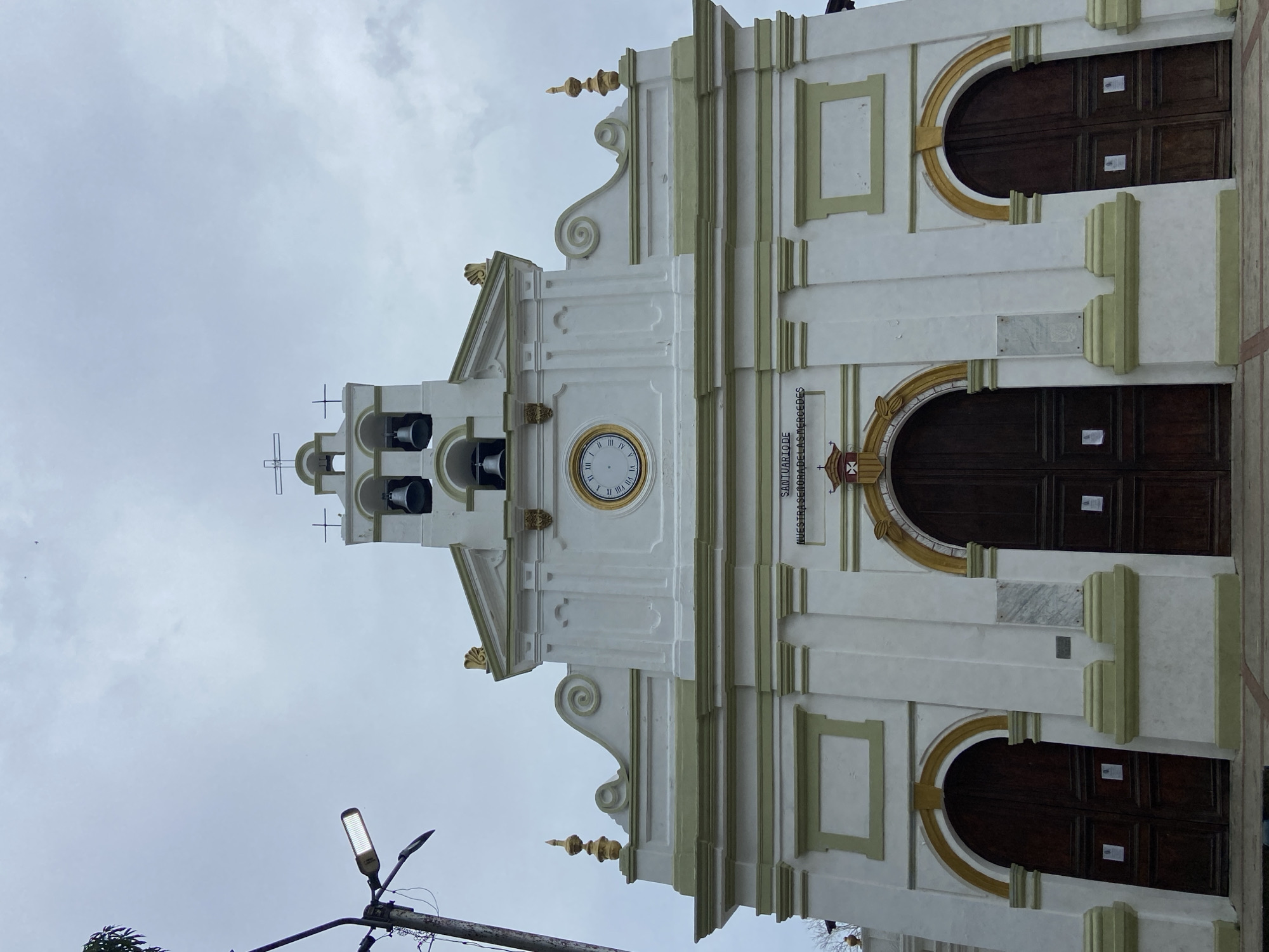 The image shows the facade of a church in Río Chico, Venezuela. The church is white with light green and gold trim. It has two large wooden doors with arched entrances. Above the doors, there is a clock and a bell tower with a cross on top. A streetlight is visible in the lower left corner. The sky is cloudy.