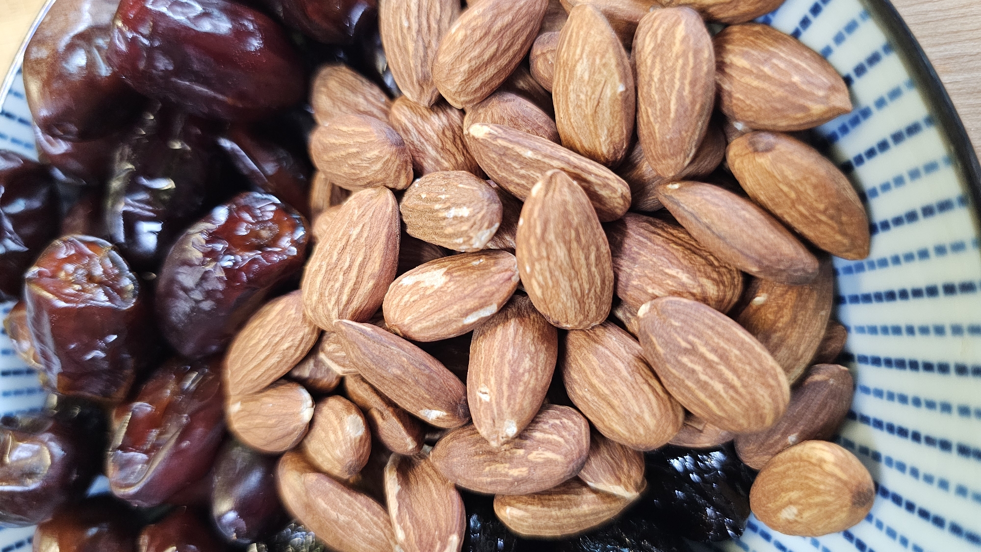 The image is a close-up, top-down view of a bowl filled with dates and almonds. The bowl has a blue and white striped pattern on its rim. The dates are dark brown and appear dried, with some showing wrinkled surfaces. The almonds are light brown, whole, and have a textured surface. They are scattered over and around the dates. The background is dark, suggesting the image was taken indoors in a controlled lighting environment. There are no people or animals present in the image. The image does not contain any information or visual cues that would allow for the determination of its location in Asan-si, South Korea.