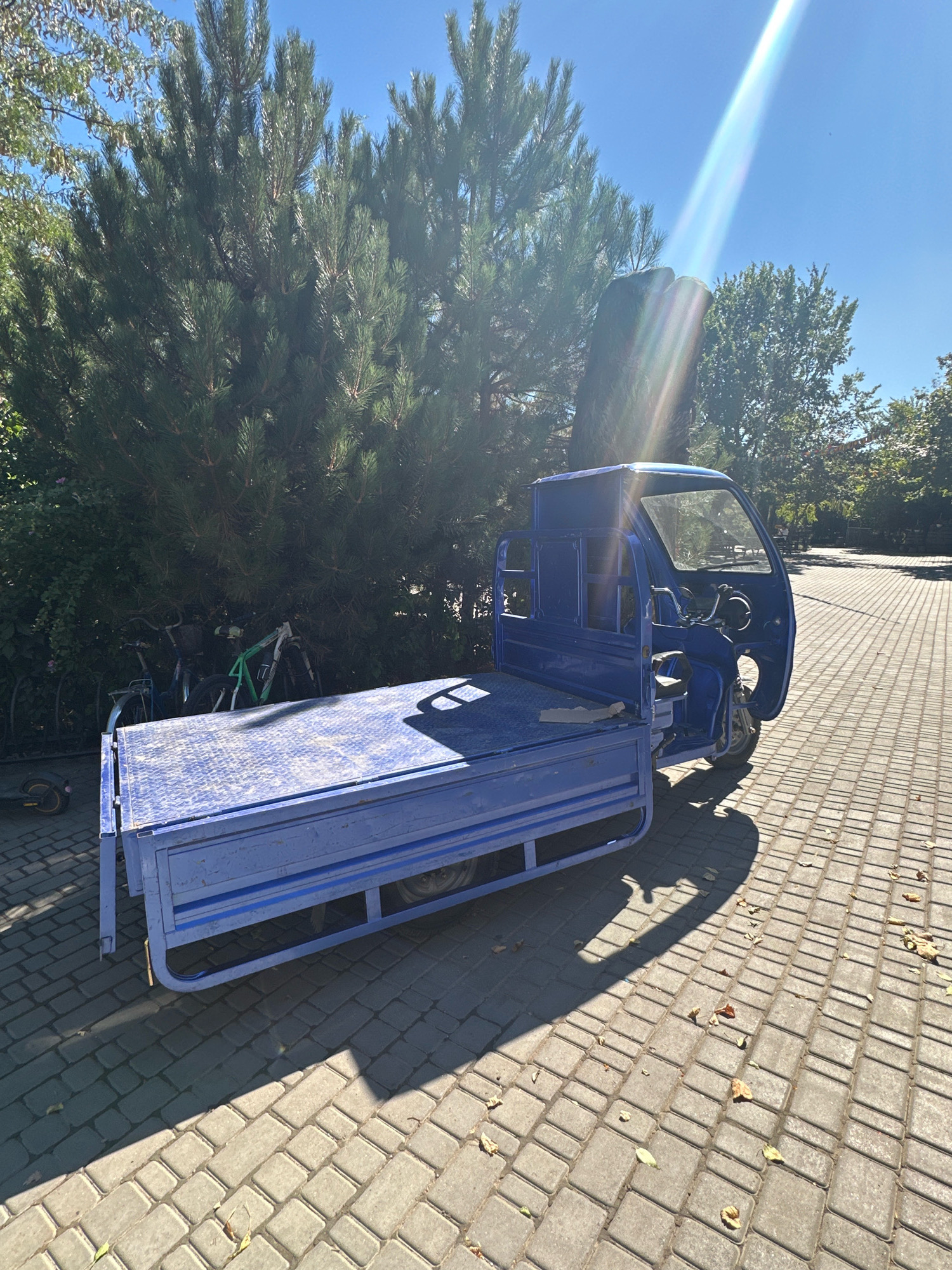 A blue, three-wheeled utility vehicle with an open cargo bed is parked on a paved area composed of rectangular gray bricks. The vehicle has a single front wheel and two rear wheels, a driver's cabin with a steering wheel and seat, and an exposed chassis. Behind the vehicle, several bicycles are leaning against a large, dense pine tree. To the right of the vehicle, the paved area extends into the distance, with trees and foliage visible on either side. The sky is bright blue with a visible sunbeam casting a lens flare effect. The scene appears to be outdoors, likely in a park or pedestrian area. The location is identified as Molodizhne, Ukraine.