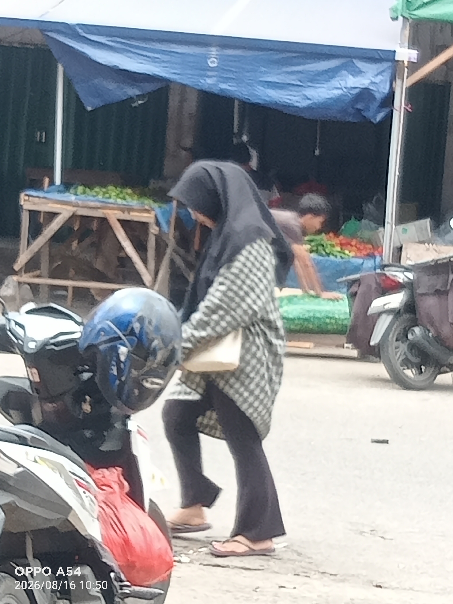 The image captures an outdoor market scene in Jambi, Indonesia, with a focus on a woman in the foreground. She is wearing a dark hijab and a checkered patterned shirt over dark pants, with her face obscured. A light-colored bag is held in her right hand. To her left, a blue helmet rests on the handlebars of a parked scooter. Behind the woman, a man is partially visible, tending to a display of fruits and vegetables, which are predominantly red and green. The market stall features a wooden structure covered by a blue tarp, and further stalls with produce are visible in the background. The ground is paved, and a second scooter is parked to the right of the frame. The scene is illuminated by natural daylight.