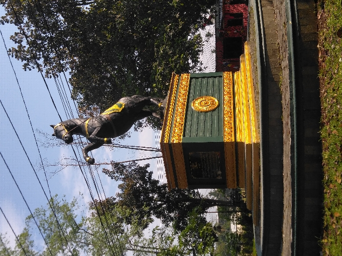 The scene depicts a large statue of a horse rearing on its hind legs, positioned on a multi-tiered pedestal, located in Hegarmanah, Indonesia. The statue, predominantly dark in color, features gold-colored bridle details and a yellow-gold saddle blanket. It is mounted on a rectangular pedestal with dark green panels and intricate gold-colored trim and ornamentation. A circular gold emblem is visible on the right side of the pedestal, and a dark rectangular plaque is present on the left side. The pedestal rests upon a multi-level, circular base constructed of dark stone or concrete. The statue and its base are situated in a grassy area with scattered dry leaves. In the background, lush green trees are visible on both sides of the statue, with some bare tree branches also present. Overhead, multiple power lines traverse the clear blue sky. To the far right in the background, a red-colored structure and other distant buildings can be discerned. The scene is illuminated by bright daylight, casting visible shadows.