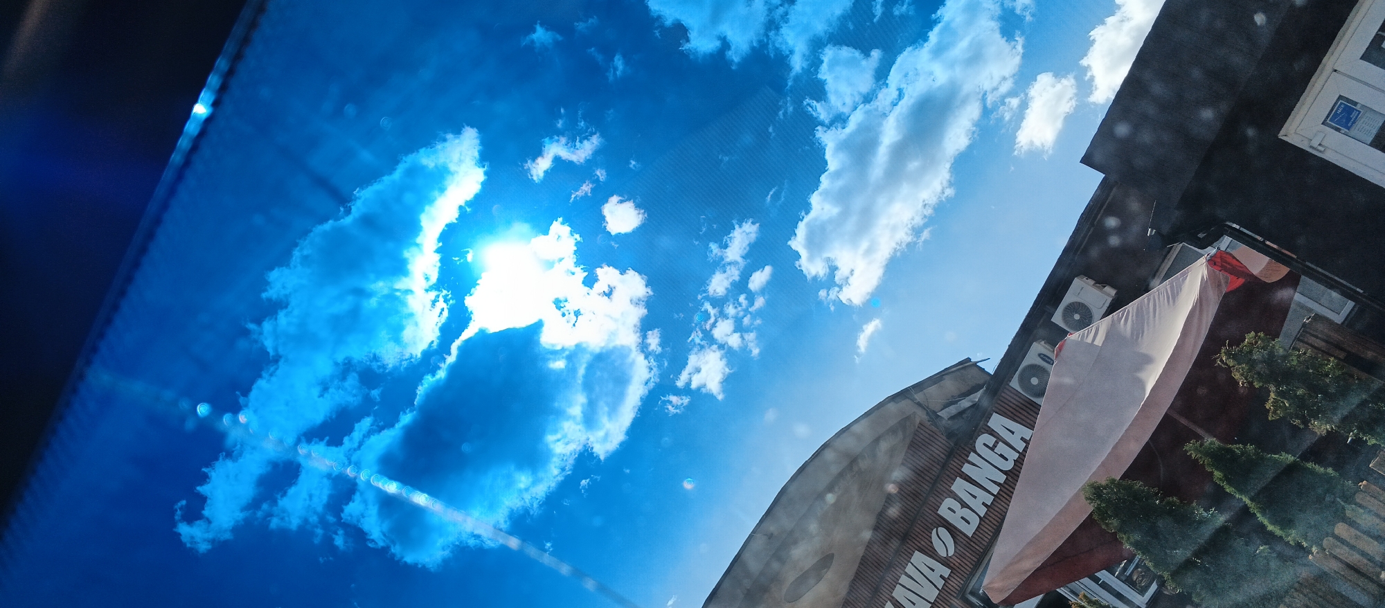 The image displays a bright blue sky filled with scattered white clouds, with the sun shining intensely and creating lens flares. Below the sky, a building is visible, angled from the bottom right corner upwards. The building has a dark roofline and features several air conditioning units mounted on its side. A large white banner with red accents is draped diagonally across the building. Text is visible on the building's facade, appearing to be part of a sign that reads "KOVA BANGA" with a coffee bean logo. The overall scene suggests an outdoor view, possibly from a window or reflective surface, given the presence of light reflections and distortions. The orientation of the image is rotated counter-clockwise.
