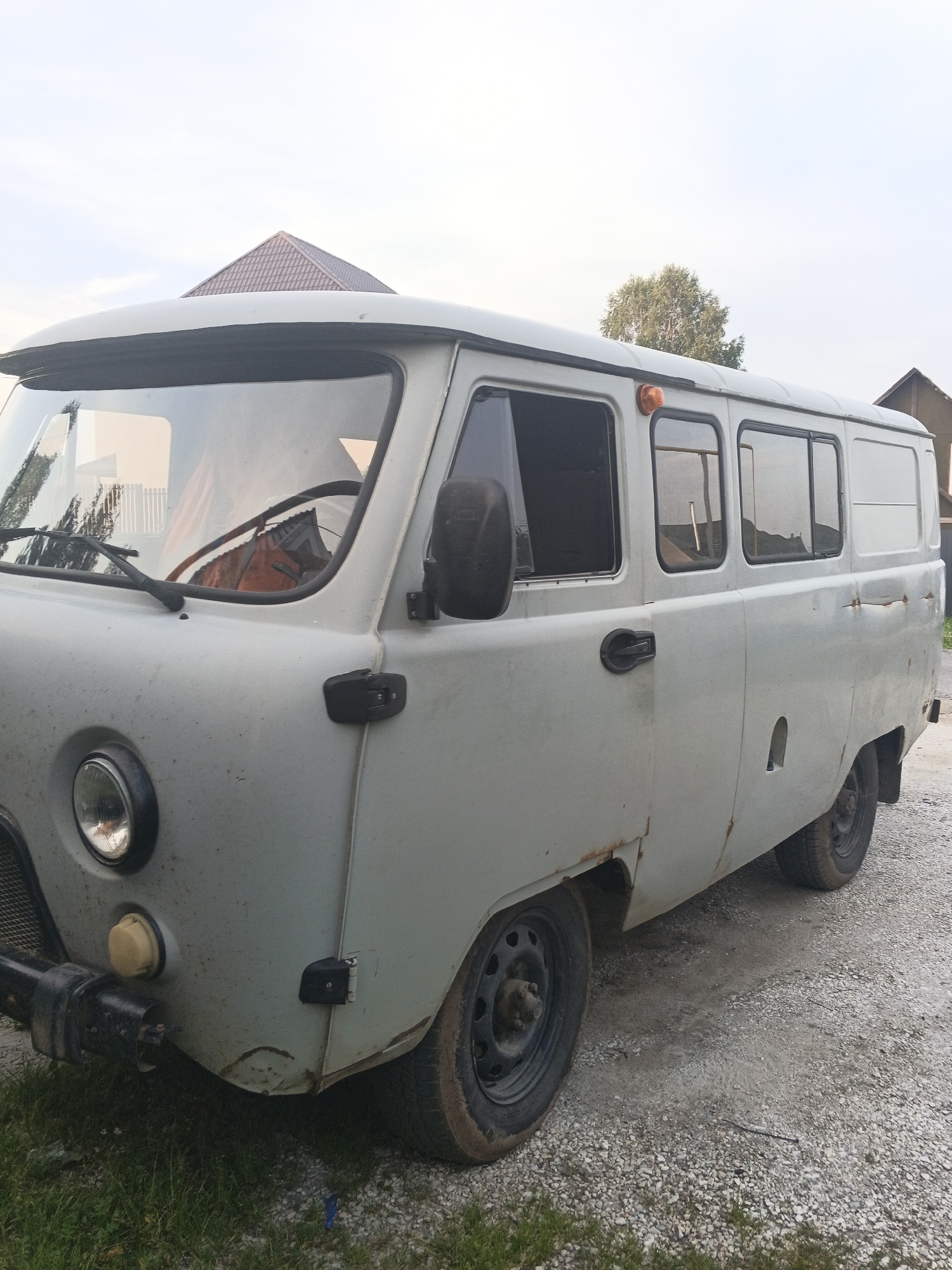 A weathered, light gray UAZ-452 van is parked outdoors on a gravel surface with some grass on the left. The van is viewed from the side, showing its front left portion prominently. The vehicle exhibits signs of age and use, with rust spots visible along the bodywork, particularly around the rear wheel arch and on the door. The van has a single round headlight on the left front, a black bumper, and black windshield wipers. There are several windows along the side, some of which are tinted. An orange turn signal is visible on the roof towards the front. In the background, a brown, gabled roof of a building is visible, along with a tree and a portion of another structure. The sky is overcast. The location is identified as Medvedyovka, Russia.