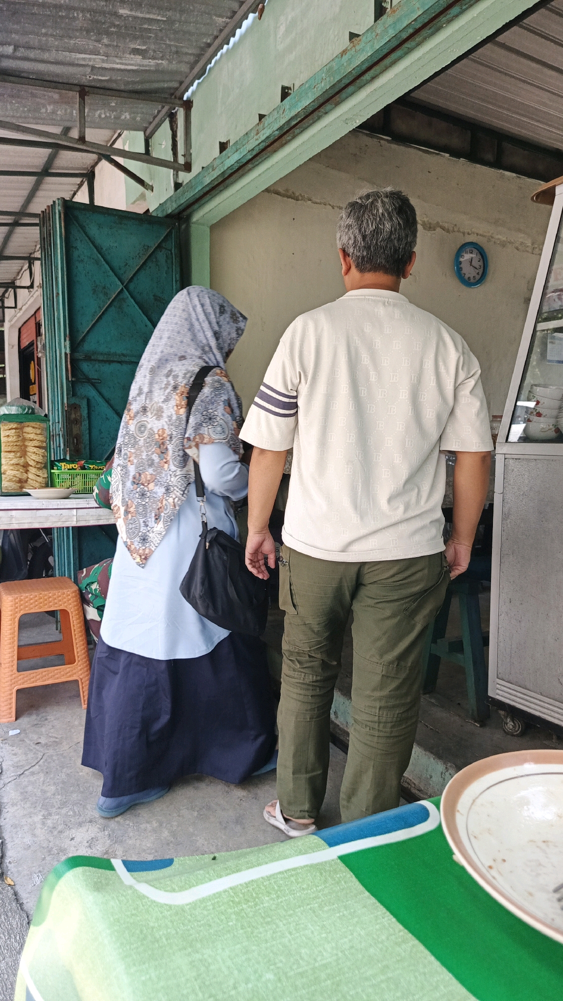 Two individuals, a woman wearing a hijab and a man, are standing outdoors in front of a building. The woman, viewed from the back and side, is wearing a blue top, a dark blue skirt, and a patterned hijab. She has a black purse slung over her shoulder. The man, also viewed from the back, is wearing a light-colored t-shirt with dark stripes on the sleeves and olive green cargo pants.
The environment appears to be a street-level establishment, possibly a food stall or small shop, with a corrugated metal roof overhead. There is a teal-colored metal door to the left and a display case or window to the right. Various items are visible on shelves and counters, including what appear to be packaged snacks. An orange plastic stool is positioned to the left of the woman. The ground is concrete. A round clock with a blue face and white numbers is mounted on the wall behind the man. In the foreground, a table covered with a green and white patterned cloth is partially visible, with a plate on it. This scene is located in the city of Surakarta, Indonesia.