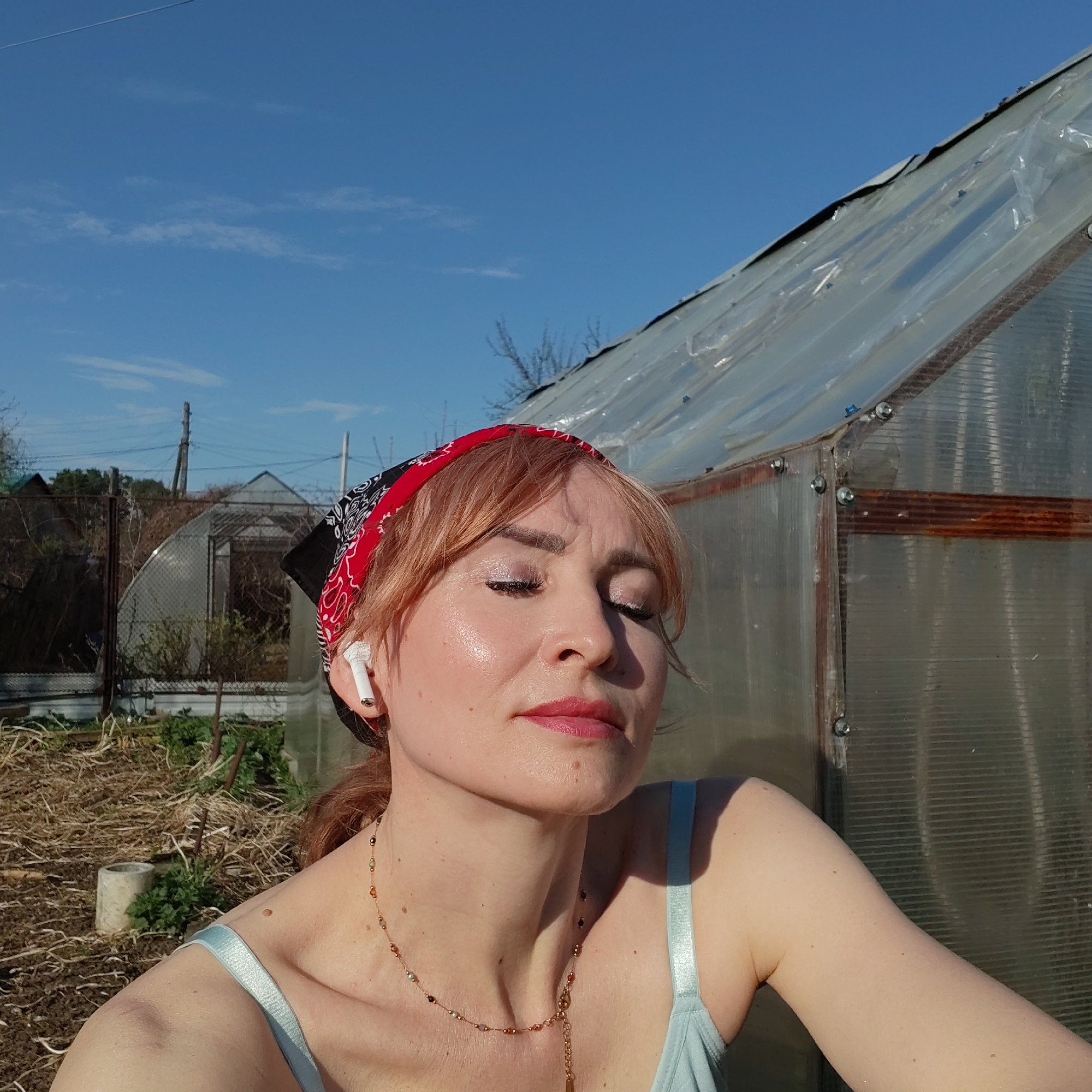 A woman with reddish-blonde hair and a red bandana is posing outdoors with her eyes closed, wearing a light blue tank top. She has fair skin and is wearing an AirPod in her left ear. She is also wearing a delicate beaded necklace. The background includes a greenhouse, a garden with visible plants, and other structures like fences and buildings, all under a clear blue sky with a few scattered clouds. The location is likely in the city of Chaikovsky, Russia.