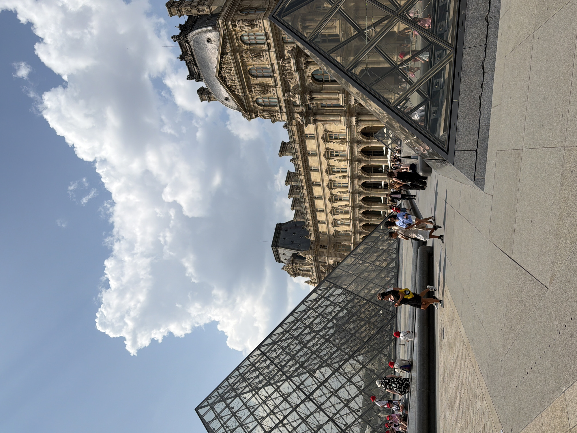 A sunny day at the Louvre Museum in Paris, France, is captured in this image. Two glass pyramids, iconic structures of the museum, dominate the foreground. The larger pyramid is on the left, its geometric facets reflecting the sky and surrounding architecture. The smaller pyramid is on the right, partially obscuring the building behind it.
The Louvre's grand facade, a classic example of French Renaissance architecture, stretches across the background. Its detailed stonework, arched windows, and ornate sculptures are visible under a bright blue sky dotted with fluffy white clouds.
Several people are scattered around the plaza, engaged in various activities. Some are walking, others are seated near the water feature in front of the larger pyramid. A group of children wearing red hats are visible in the lower left corner. The overall scene conveys a sense of a bustling tourist destination on a pleasant day.
