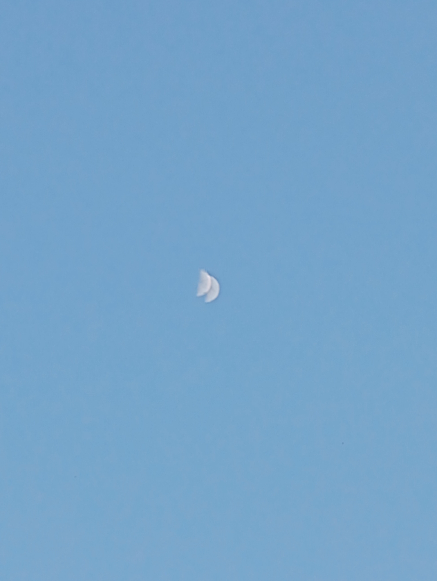 A distant, crescent-shaped moon is visible in a clear, pale blue sky. The moon appears small and is positioned slightly to the left of the center of the image. The sky is devoid of clouds, and there are no other celestial bodies or objects in view. The overall impression is one of vast, empty space.