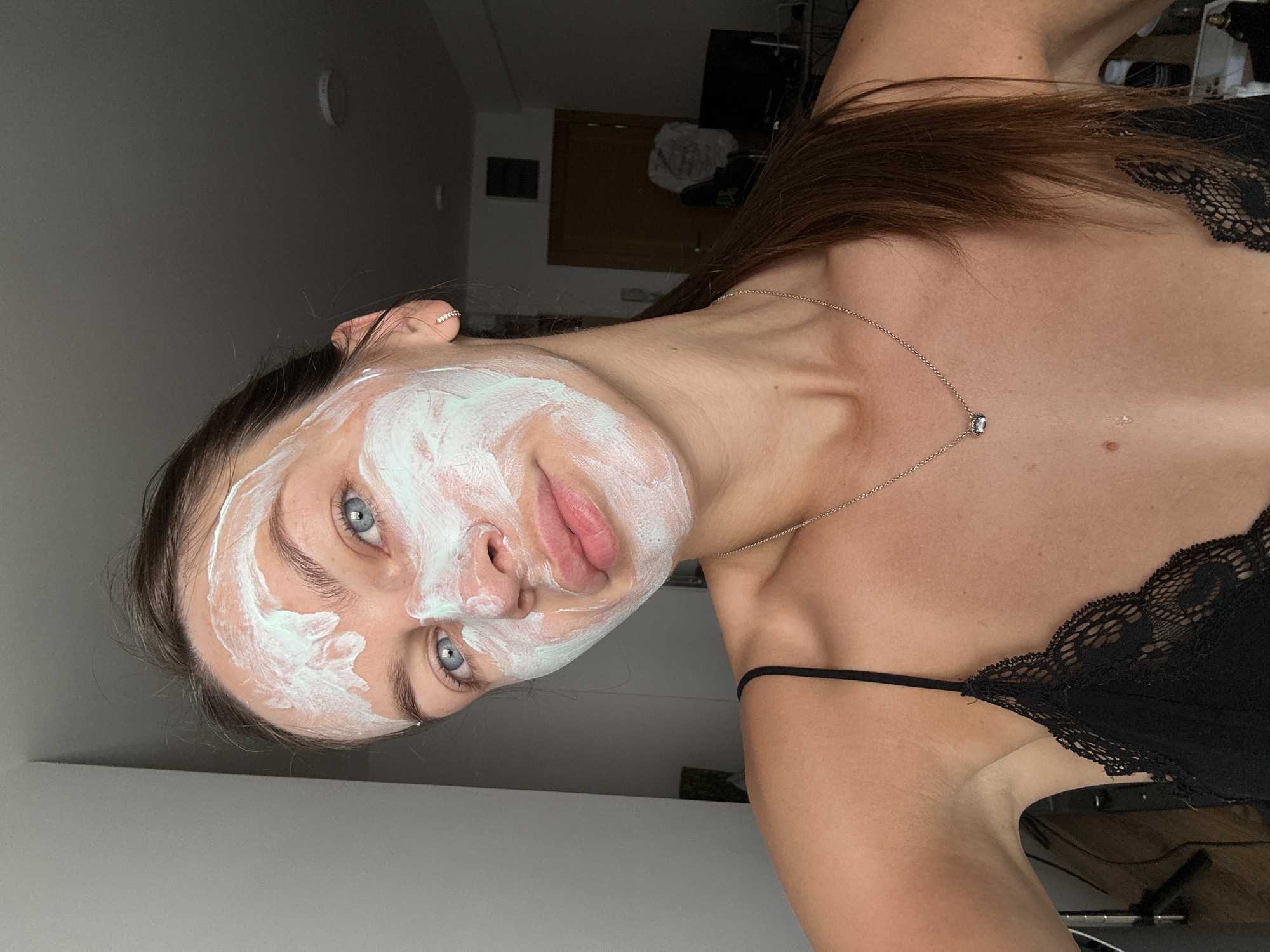 A young woman with blue eyes and brown hair pulled back in a ponytail is applying a white and green face mask. She is wearing a black lace bralette and a silver necklace with a small charm. The background is a dimly lit room with a gray wall on the left and a doorway and furniture visible on the right. The scene is indoors. The image was captured in Donji Pobori, Montenegro.