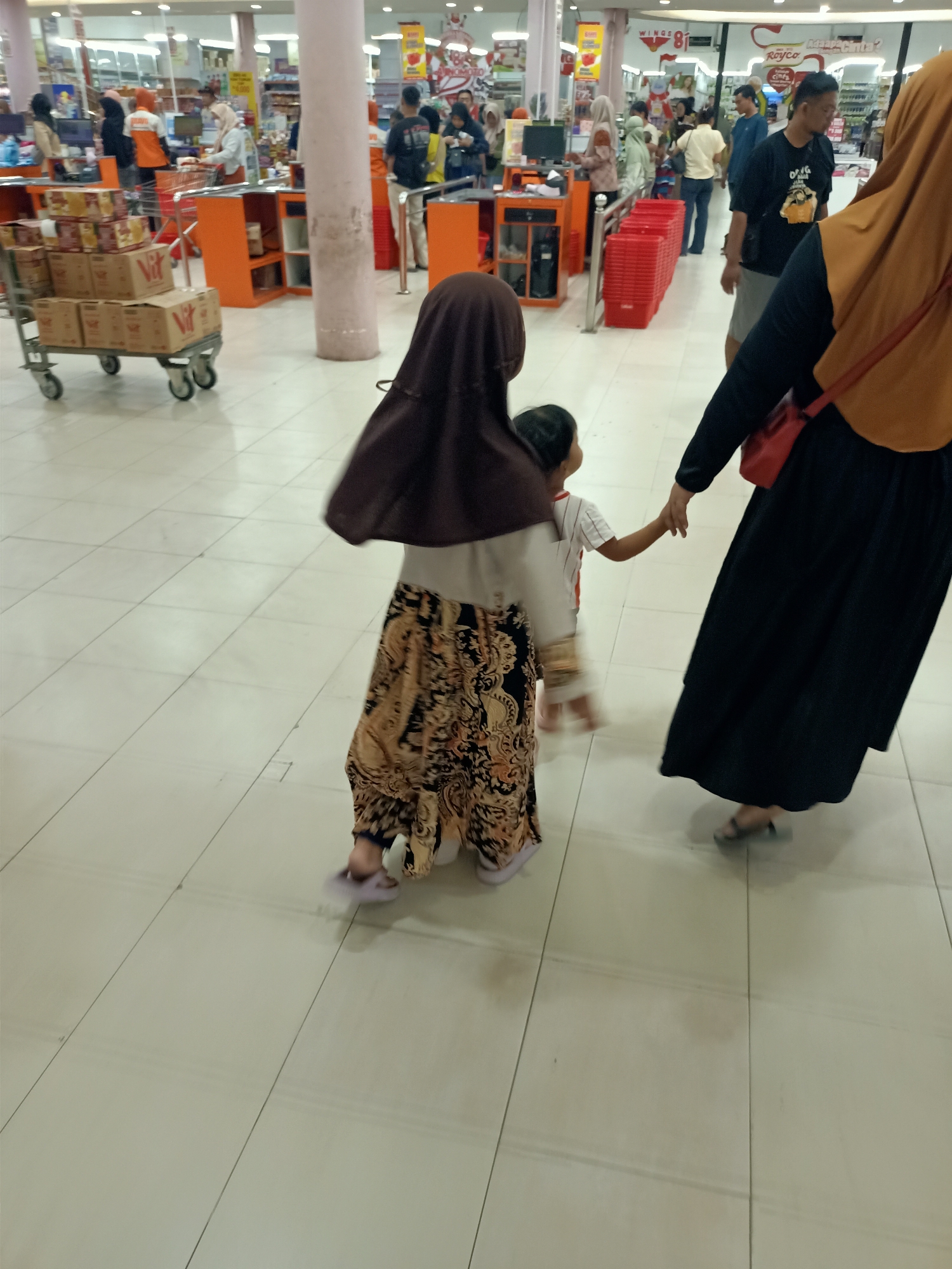 This image depicts a scene inside a supermarket in Mernung, Indonesia. A group of people, including adults and children, are seen walking through the aisles. In the foreground, a woman wearing a dark hijab and a black abaya, with an orange scarf draped over her shoulder, holds the hand of a young child. The child, wearing a white t-shirt with red stripes, is also holding the hand of another child, who is partially obscured but appears to be wearing a brown hijab and a patterned skirt. In the background, other shoppers and store employees are visible, along with shelves stocked with products and checkout counters. The environment appears to be a typical indoor retail space with tiled flooring and fluorescent lighting.