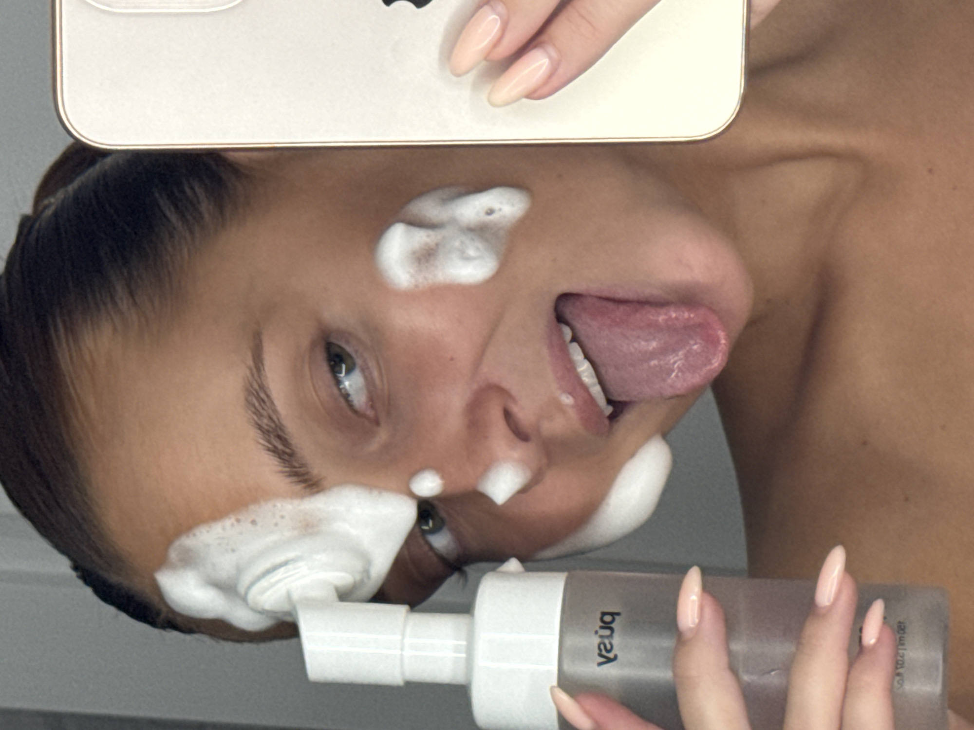 In a close-up, self-taken photograph, a young woman is captured with her face partially covered in white foam, suggesting a facial cleansing routine. She holds a clear bottle of cleanser in her left hand, with the brand name "push" visible in black text. Her tongue is extended, and her eyes are looking towards the camera, which is being held in her right hand, partially obscuring her face. She is wearing light-colored, oval-shaped fingernails. The background appears to be indoors, with a neutral-colored wall. There is no discernible indication of the city of Chaikovsky, Russia, or any urban environmental elements present in the image.