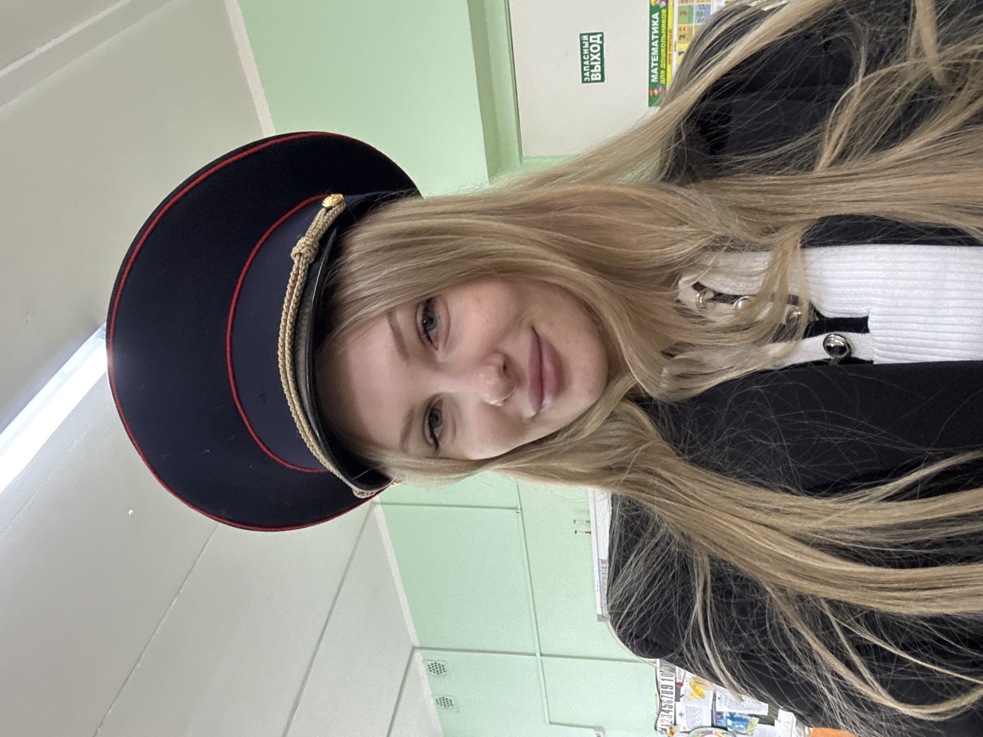 A young woman with long blonde hair, wearing a black jacket over a white and black shirt, and a black and red peaked cap with gold trim, is posing indoors. The cap resembles a police or military-style hat. The background is a classroom or similar institutional setting, with light green walls, a door with "Запасный Выход" (Emergency Exit) signage in Russian, and a poster with numbers and math-related information, likely for children. The picture appears to have been taken in Perm, Russia.