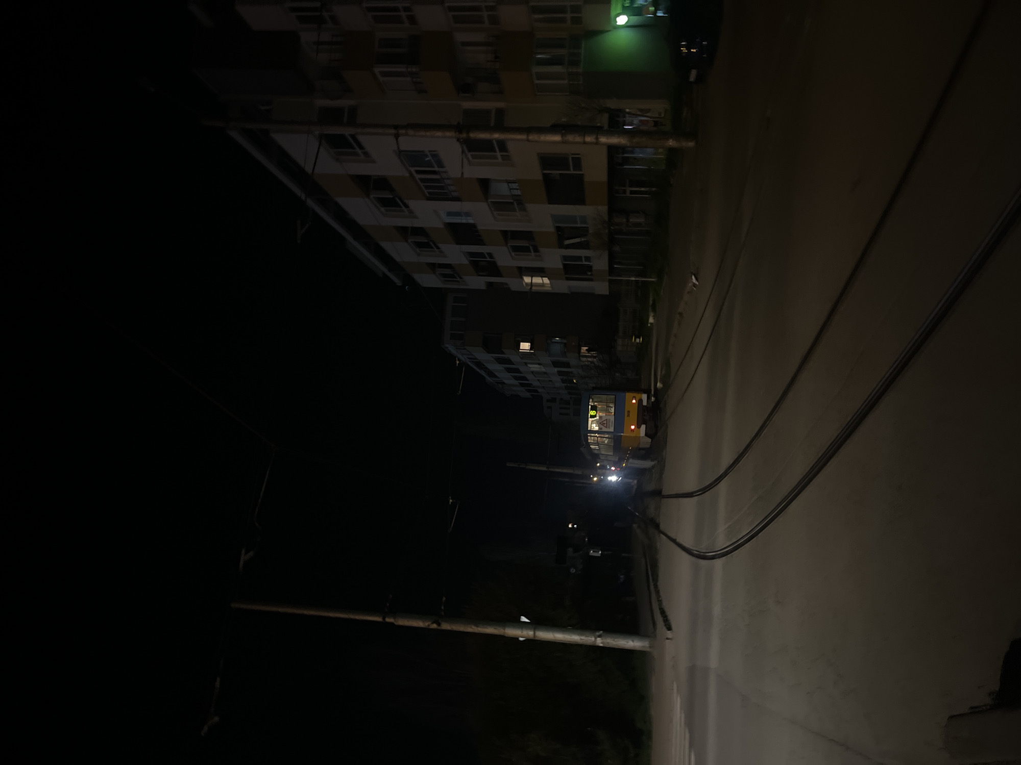 At night in Sofia, Bulgaria, a tall building with lit windows stands on one side of a street. Power lines run alongside the road. Further down the street, a tram is visible. The scene is dimly lit.