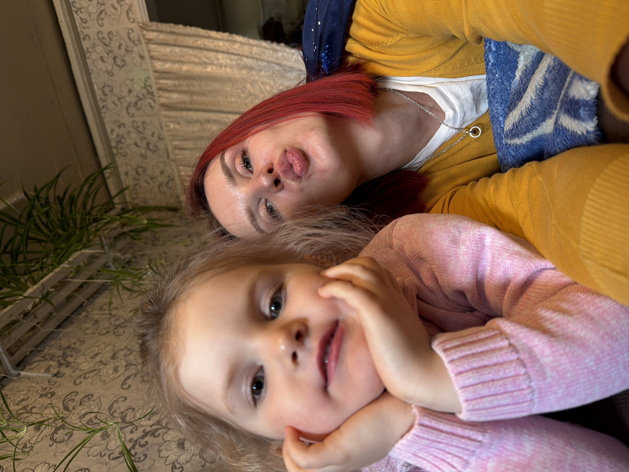 This intimate, low-angle photograph captures a playful moment between an adult and a young child, likely a mother and her daughter, in an indoor setting in Заболотово, Russia. Both subjects are positioned as if lying down, looking towards the camera.
The adult, located in the upper portion of the frame, has distinctive short, bright red hair. She is seen making a whimsical "kiss face" with pursed lips and wide eyes, conveying a sense of lightheartedness. She wears a yellow knitted cardigan over a white top, accessorized with a silver chain necklace featuring a circular pendant with a dark brown center. A dark blue fabric with white speckles, possibly a scarf or blanket, is draped behind her head.
Below and slightly in front of the adult is the young child, who appears to be a toddler or preschooler. She has light blonde or light brown hair and a soft, sweet expression, with her hands gently resting on her cheeks. She is dressed in a cozy pink knitted sweater.
The background features a light-colored surface, possibly a wall or upholstered furniture, adorned with an elegant, swirling floral pattern in a muted grey or beige tone. To the left, a white wooden lattice structure is partially visible, along with some green leafy plants, suggesting indoor greenery. A blue and white striped or patterned blanket is visible between the two individuals. The lighting is soft and ambient, characteristic of an indoor daytime setting, enhancing the warm and familial atmosphere of the scene. There is no visible text in the image.