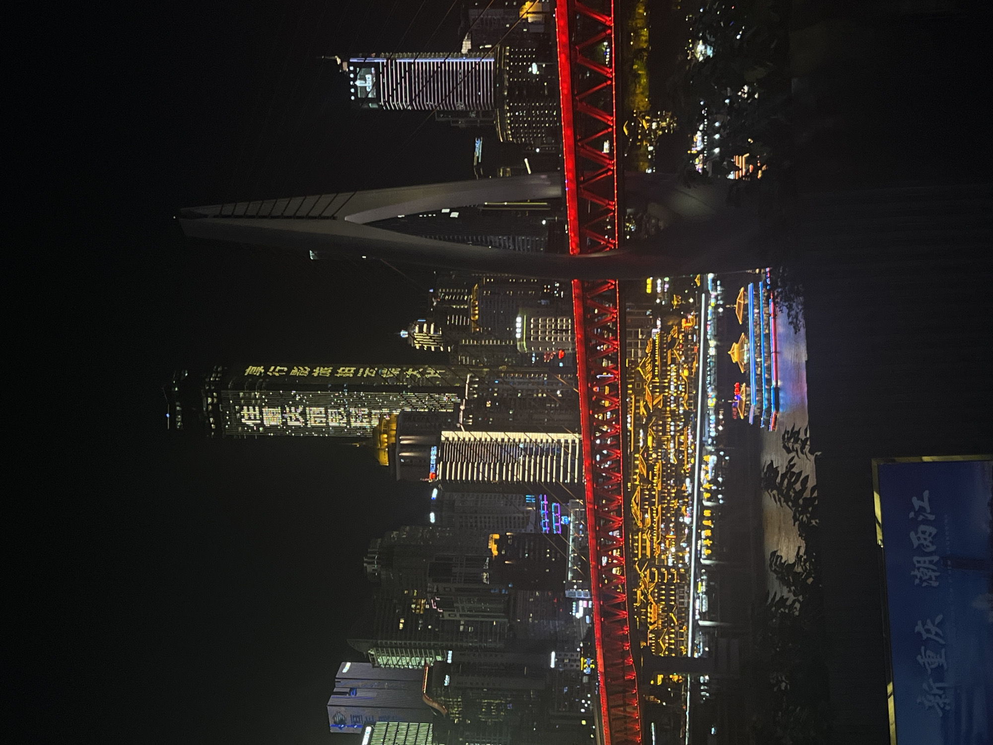 This is a nighttime photograph of the Chongqing skyline, specifically showing the area around Jiangbeicheng Street. The image features a waterfront with a brightly lit bridge spanning across a body of water. The bridge is illuminated with red lights. Along the waterfront, there are many buildings with traditional Chinese architecture, also lit up with warm yellow lights.
In the distance, modern skyscrapers rise above the traditional buildings, some of which are adorned with vertical lines of white and blue lights. One tall building on the left side of the image displays yellow text vertically, likely a sign or advertisement. A large, distinctive cable-stayed bridge structure is prominently visible in the center-right of the frame.
A boat with multiple decks and colorful lights, including blue and red accents, is on the water, likely a passenger vessel. In the foreground, silhouetted foliage obscures the lower part of the scene, with a sign in the bottom left corner displaying Chinese characters, possibly indicating the location "新重庆 潮两江" (New Chongqing, Facing the Two Rivers). The overall scene depicts a vibrant urban waterfront at night.