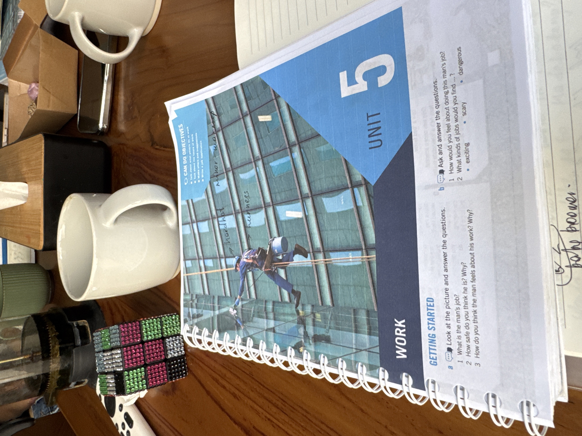 The image shows a book titled "Unit 5: Work" with a picture of a window cleaner on the cover. The text includes discussion questions about the safety and feelings associated with the job. The book is open on a table with several objects: two white mugs, a box, a tissue dispenser, a coffee press, and a bejeweled cube. Notes are visible in the margins of the open pages. The location, Kuta Selatan, Indonesia is not evident.