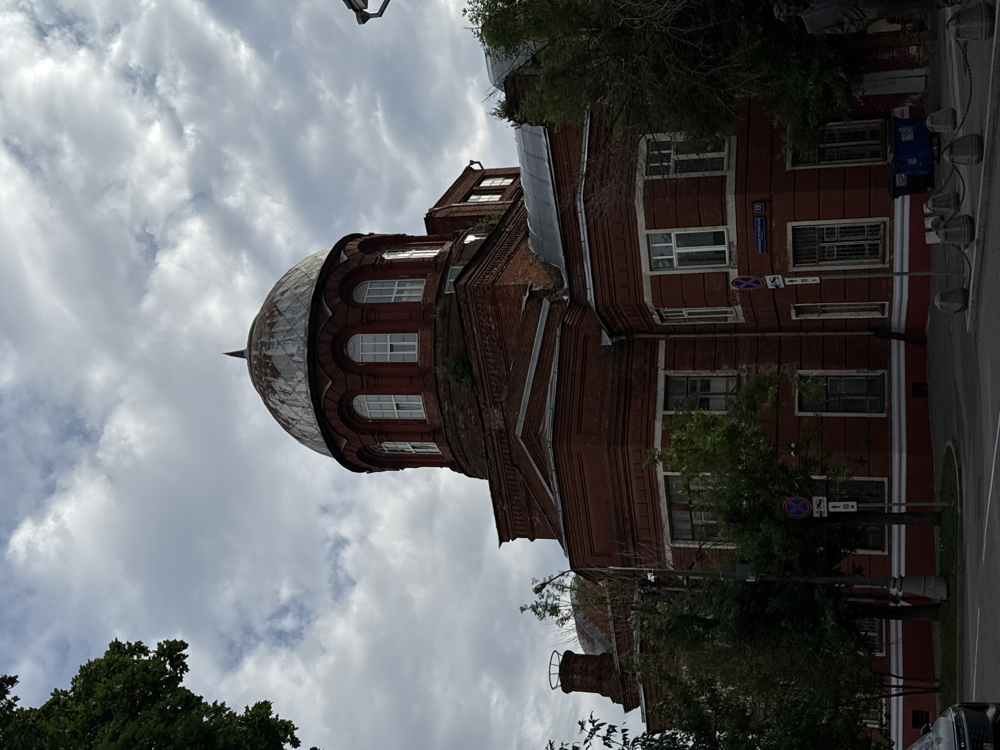 The image shows the red brick building of the former Church of the Archangel Gabriel in Moscow, Russia, located at 13 Chistye Prudy Boulevard. The building features a large, weathered dome with arched windows. There are trees around the building, and a cloudy sky is visible. In the foreground, there is a street with a sidewalk and some bollards. A dark-colored car is partially visible on the left side of the image. The building's facade is intricate, with layered brickwork and white-trimmed windows. A street sign with Russian text and the number 13 is mounted on the corner of the building.