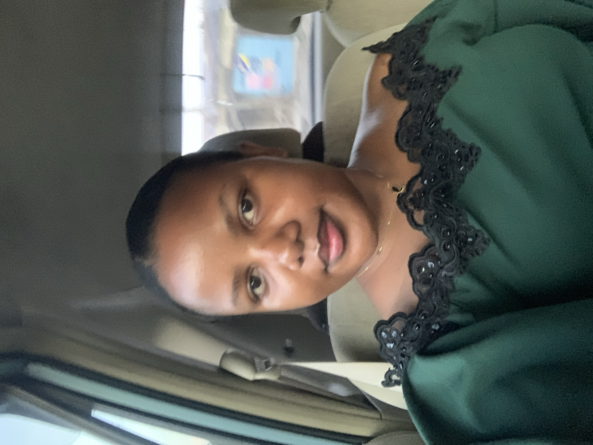 The image shows a woman in a car. She is wearing a dark green dress with black lace trim around the neckline. She is also wearing a gold chain necklace. The car appears to be moving through a city. In the background, signs can be seen, likely indicating the location is in Dar es-Salaam, Tanzania. The car's interior is light-colored.
