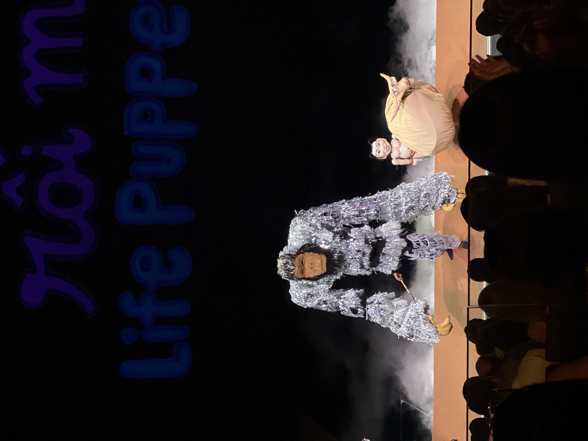 The image depicts a theatrical performance in Bac Nha Trang Ward, Vietnam. Two large puppets are center stage, viewed from the audience. One puppet, dressed in a shimmering silver costume, is holding a long object. The second puppet appears smaller, dressed in beige, with an exposed midriff. The audience is visible on the right side of the image. A sign reading "Life Puppe" is present on the left side of the image. White smoke or fog is present on stage, obscuring the back of the stage.