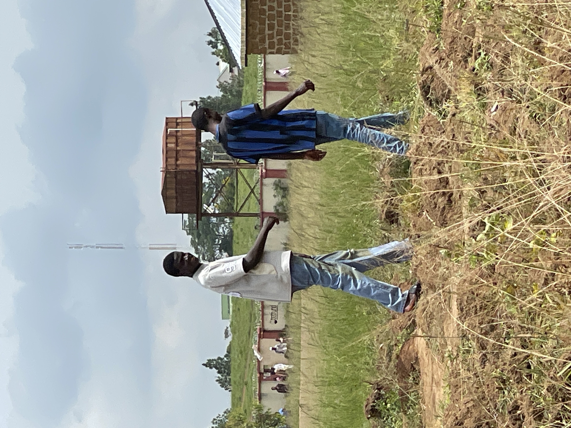 The media file shows a rural outdoor scene in Durumi, Nigeria. Two men are the primary subjects, standing in a grassy, somewhat overgrown area. The man in the foreground wears a light-colored short-sleeved shirt and blue jeans. He appears to be looking upwards. Behind him and slightly to the left, another man is visible, wearing a blue and black striped t-shirt and blue jeans. He is also looking upwards and to the side, with his arm extended.
In the background, a building with a red-brick section and a white roof is present. Adjacent to it, a tall structure with a water tank on top is visible, supported by metal scaffolding. Further in the background, more buildings and trees can be seen. A few other people are visible near the building, and some animals, possibly goats or sheep, are also present.
The ground in the foreground is a mix of dry grass and soil, suggesting a potentially arid or dry season. The sky is overcast with clouds, indicating a possibly cloudy or impending rainy day. The overall impression is one of everyday rural life. No visible text is discernible in the image. The lighting suggests it is daytime, likely late morning or afternoon.