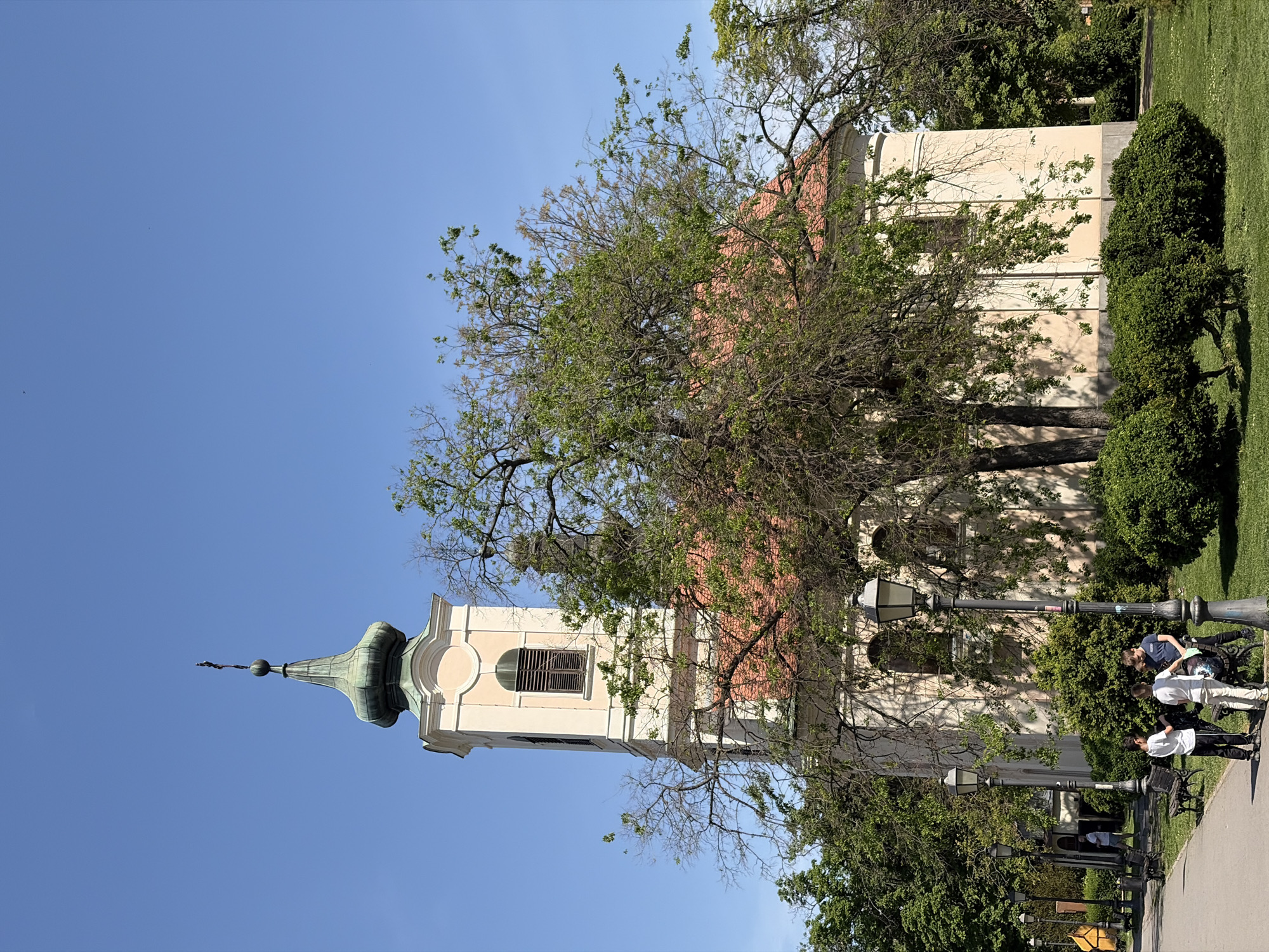 A brightly lit, daytime photograph captures a historic church tower and a park setting in the Zemun Urban Municipality, Serbia. The central feature is a tall, light-colored church tower with an ornate green, onion-domed spire topped by a finial or cross. Behind the tower, part of a larger building with a reddish-orange tiled roof and light-toned walls is visible, partially obscured by trees.
The scene depicts an urban park or garden, characterized by numerous green trees with varied foliage, some appearing dense and leafy, others showing more bare branches. Manicured green grass sections are present to the right of the building. A paved pathway runs through the lower right portion of the image, bordered by several traditional black lampposts and a park bench.
At least three young individuals are visible near the pathway and lampposts. One person is seated on the bench, another leans against it, and a third stands nearby. They are casually dressed and appear to be relaxing or socializing in the park.
The weather is clear and sunny, indicated by the vibrant blue sky with no visible clouds and strong shadows cast by trees and structures, suggesting it is either late morning or early afternoon. No discernible text is visible on any objects or signs within the frame. The overall atmosphere is calm, blending historical architecture with natural elements in an urban recreational space.