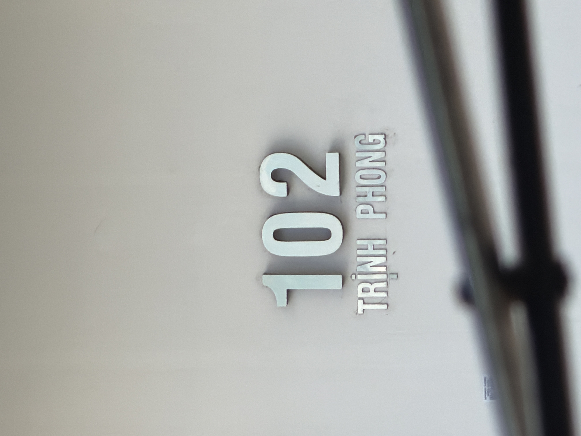 A building facade in Nha Trang Ward, Vietnam, displays the number "102" in large, raised, metallic digits. Below the number, the word "TRINH PHONG" is also displayed in raised, metallic letters. The building's wall is a light, neutral color. In the foreground, out of focus, are two horizontal metal railings.