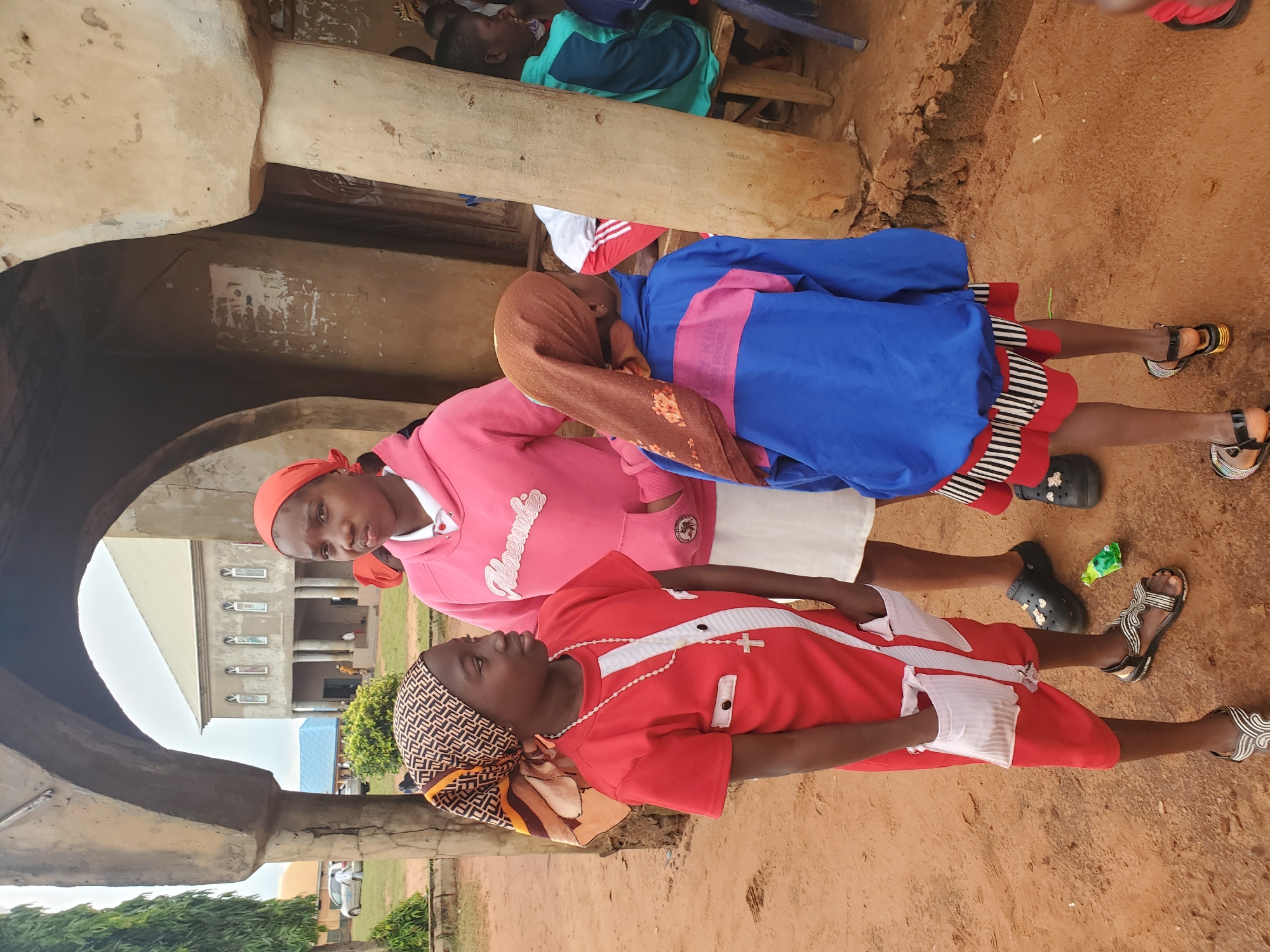 In the city of Nsugbe, Nigeria, three young girls stand in front of an arched structure with rough, textured walls. The girl on the left, wearing a red dress with white accents and a patterned headscarf, has a white beaded necklace with a cross. To her right, a girl in a pink hooded sweatshirt and a red headscarf looks towards the camera. The third girl, on the far right, is seen from the back, wearing a blue jacket with pink accents over a red and black striped skirt, and a brown headscarf. In the background, a building with columns and arched windows is visible, possibly a church. The ground is sandy and reddish-brown.