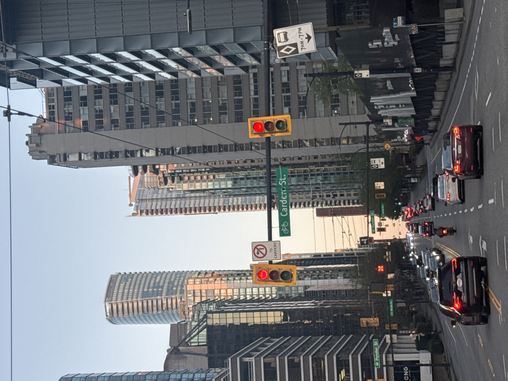 This media file captures an urban street scene in Vancouver, Canada, likely during the late afternoon or early evening, given the soft, warm light reflecting off the buildings. The image is taken from a street-level perspective, looking down a multi-lane city road lined with tall buildings.
The scene is dominated by a bustling cityscape. Numerous modern high-rise and mid-rise buildings with varied architectural styles, including many glass facades, flank both sides of the street. Reflections are prominent in the windows and surfaces of these structures. The sky appears clear and bright above the buildings.
On the street, a line of vehicles, including cars and at least one motorcycle, are visible. The traffic appears to be stopped or moving slowly, with several vehicles displaying illuminated red brake lights. A prominent traffic light in the foreground shows a solid red signal. Utility poles and overhead wires crisscross the frame.
Several signs with visible text provide location and traffic information:
* A white rectangular sign with a black border indicates "7 AM - 7 PM" with a diamond symbol and a right-pointing arrow, likely designating a restricted lane.
* A green street sign reads "Cardero St." and includes a bicycle symbol.
* Another sign shows a "No Left Turn" symbol with the text "TO CARDERO" below it.
* Further down the road, a green street sign identifies "W Pender St."
* Other distant signs, partially obscured, appear to indicate "W Hastings St" and "W Georgia St."
* Part of a "WRONG" sign is faintly visible on a pole further down the street.
* The letters "ONG" are visible on a building facade near the bottom right.