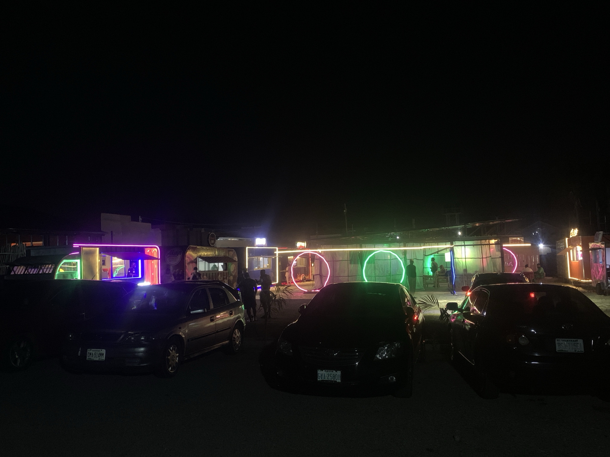 The image shows an outdoor nighttime scene in Jos, Nigeria. A row of parked cars fills the foreground. In the background, there are several food trucks or small stalls illuminated with neon lights, including circular neon displays. People are seen standing and interacting around the stalls. The environment is dark with limited ambient lighting.