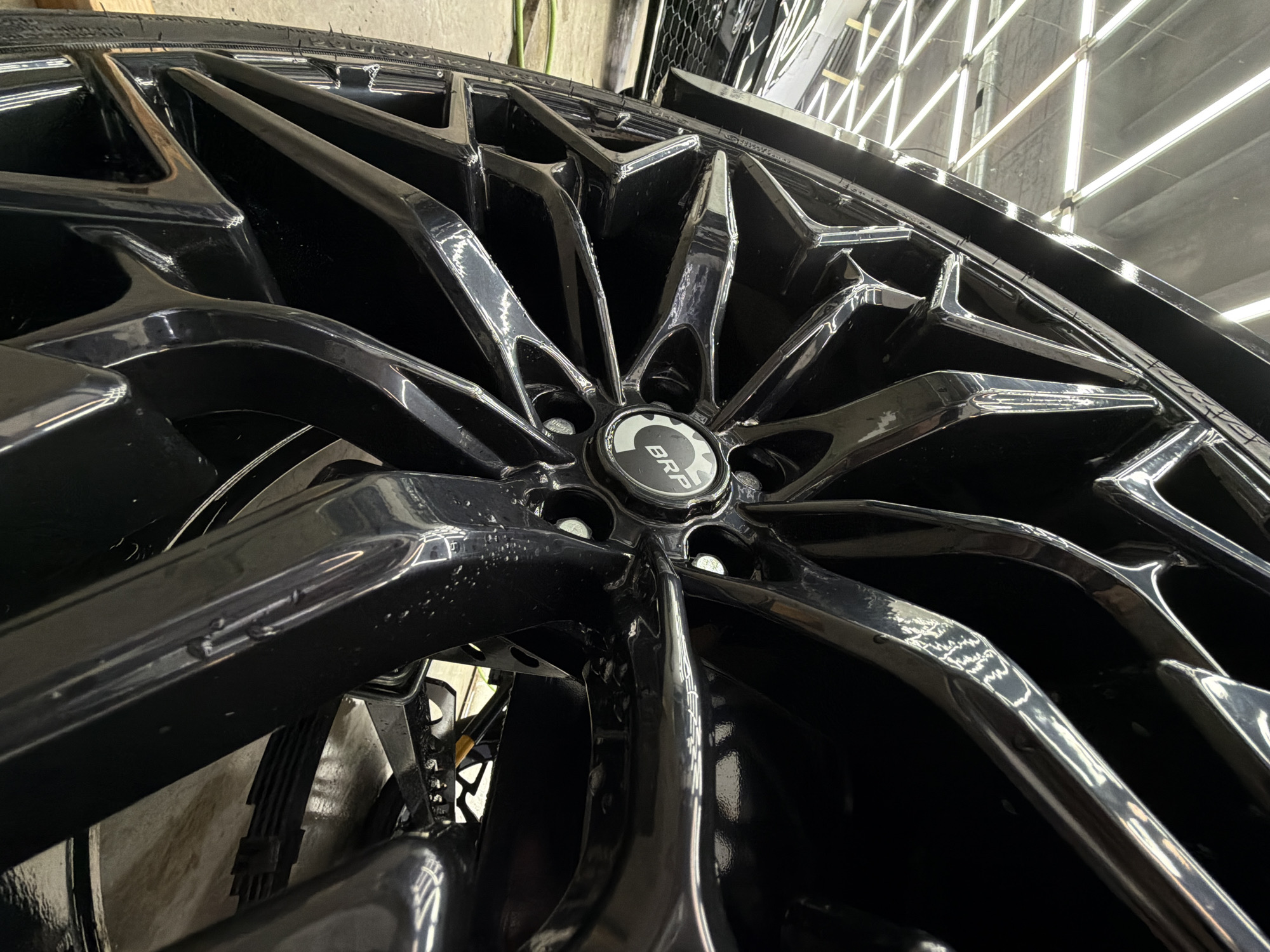 The image shows a close-up view of a black car wheel with a complex, multi-spoke design. The center cap displays a logo, possibly "BRP". The wheel appears to be clean and reflective, suggesting it may have been recently detailed or is of high quality. The background includes a wall with bright, linear lighting fixtures, indicating the scene is indoors, possibly in a garage or auto shop. I have no knowledge of the location being in Krasnoyarsk, Russia.