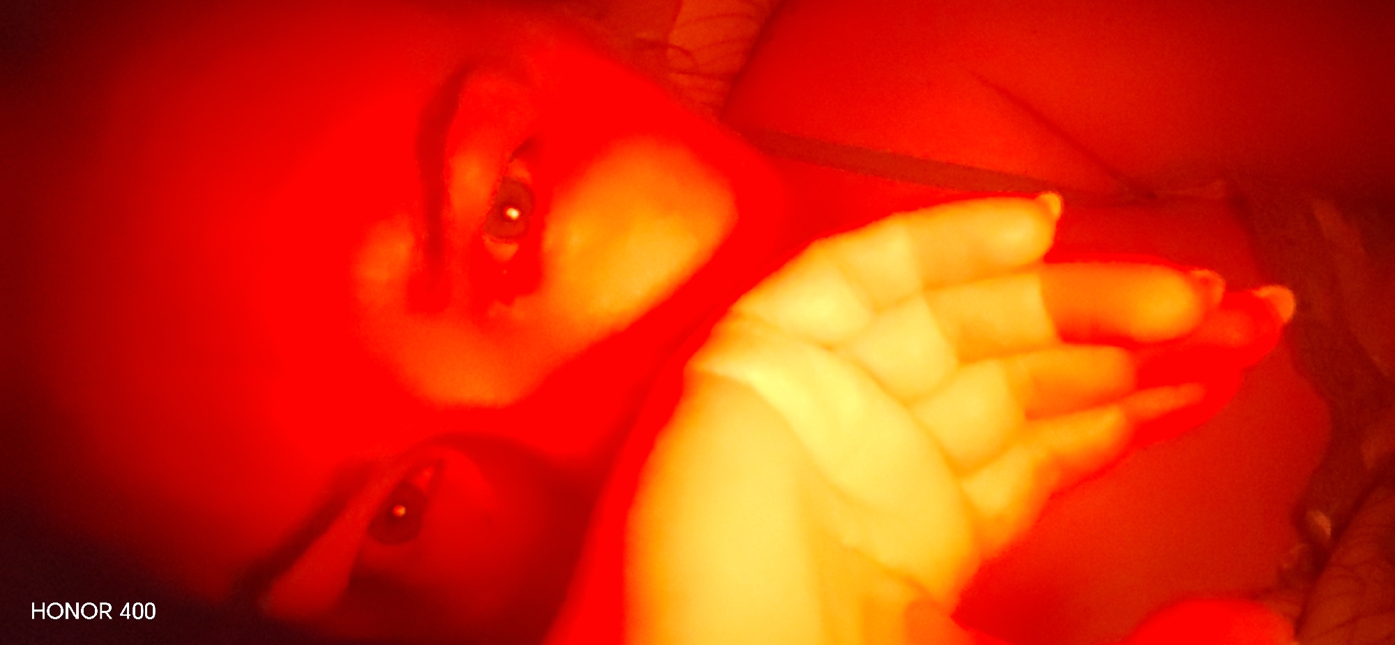 This media file presents a close-up, high-contrast image of a person, likely a woman, under intense artificial red and orange lighting. The subject is seen from the chest up, positioned at an angle as if reclining or lying down, with their gaze directed towards the viewer. Their eyes are prominent and reflective, catching the light. One hand is visible in the foreground, positioned near the face or chest, with fingers spread slightly; nail polish is discernible on some fingernails.
The scene is dominated by a vivid color palette of deep reds and warm oranges, which saturate the entire frame and cast the person's skin in glowing, luminous tones. The background is indistinct due to the extreme lighting, suggesting an indoor setting. The intense, warm illumination creates a dramatic and captivating atmosphere.
In the bottom left corner, the text "HONOR 400" is clearly visible, indicating a camera or device used. No other specific location cues or time of day can be determined from the visual content. The activity appears to be posing for a photograph.