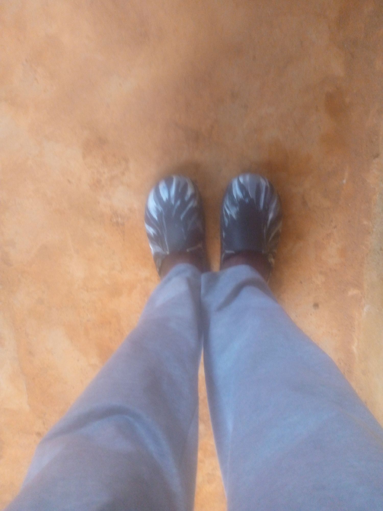 The image shows a view of a person's feet and lower legs. The person is wearing light gray sweatpants and dark-colored slip-on shoes with a white pattern. The background is a solid tan or orange color, possibly the floor or ground. The location is unknown.