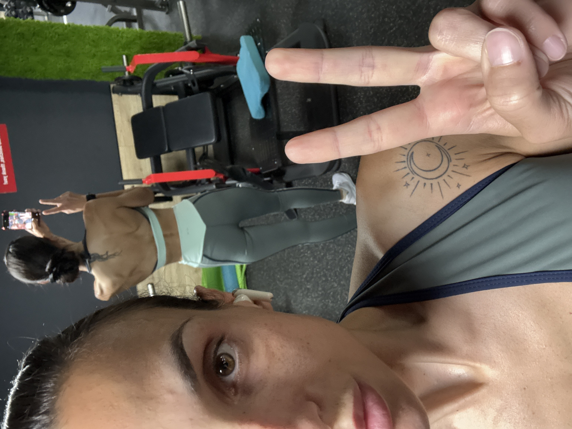The image shows a selfie taken by a woman in a gym. She is wearing a sports bra and leggings. She has a tattoo of a sun and moon on her shoulder and is making a peace sign with her hand. The gym equipment is visible in the background. There is no information to determine if the location is Pleven, Bulgaria.