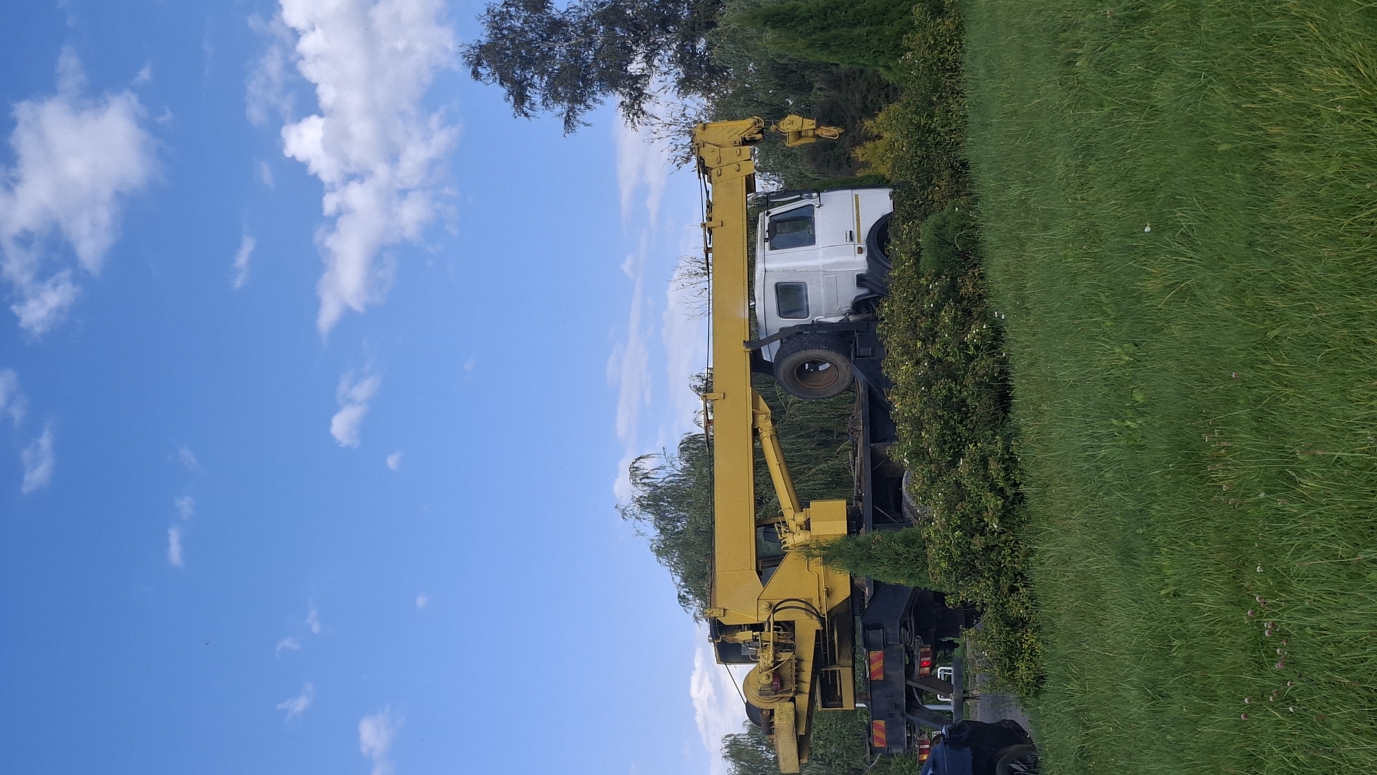 A yellow crane truck is parked in a grassy area with trees and bushes surrounding it. The truck has a long boom extended horizontally. A white cab sits on the truck chassis. The sky is blue with scattered white clouds. The scene appears to be outdoors in a non-urban setting, possibly a park or a rural area, in Myadzyel, Belarus. There are no people visible in the image.