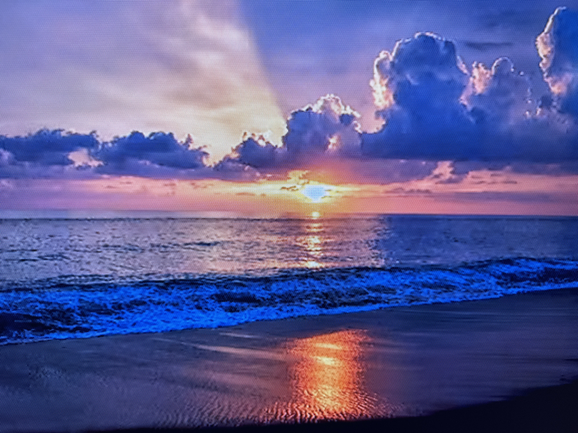 The image depicts a vibrant sunset over a body of water, likely an ocean or large lake, with a beach in the foreground. The sky is a gradient of colors, transitioning from deep blues at the top to shades of purple, pink, and orange as it nears the horizon. Dramatic clouds, illuminated by the setting sun, are scattered across the sky, with rays of light breaking through them. The water reflects the colors of the sky, creating a shimmering effect, and a line of breaking waves is visible closer to the shore. The beach appears wet and dark, with reflections of the sunset visible on its surface. There are no people, buildings, or specific landmarks visible in the image that would help to identify a precise location within Szczecin, Poland, or any other city. The scene is a natural landscape at sunset.