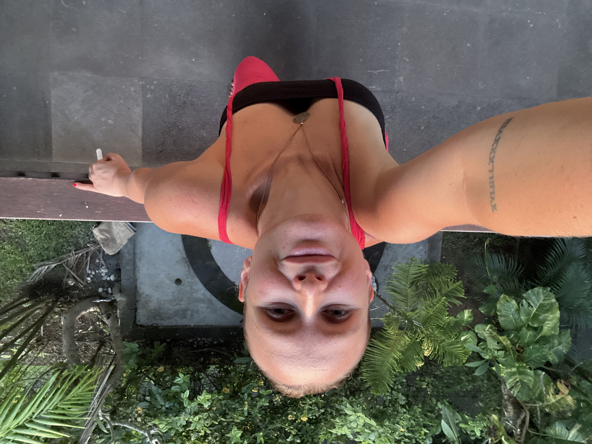 A woman stands in what appears to be an outdoor area, possibly a balcony or patio, based on the presence of a gray tiled floor and a wooden railing. She is wearing a black top with red straps and holding a white object, possibly a cigarette or chalk, in her left hand. A tattoo is visible on her upper arm. The background features tropical vegetation, including palm trees and various green plants. Below the wooden railing, there's a small garden area and a circular design element on the ground. The image's upward angle suggests the photo was taken from a higher vantage point, looking down. The location, Kuta Selatan, Indonesia, cannot be verified solely from the image. No specific actions or interactions are discernible beyond the woman holding the white object.