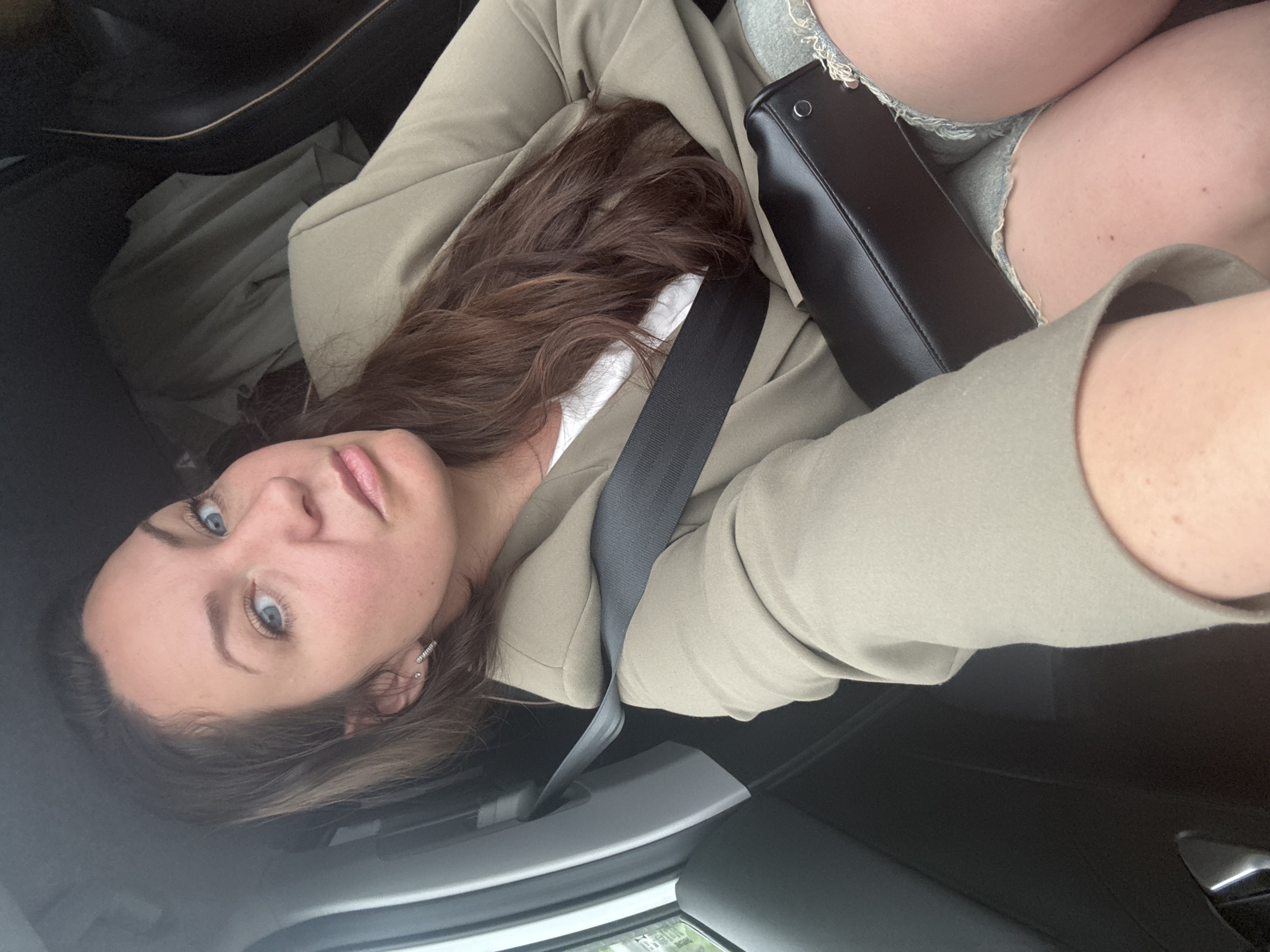 A young woman is captured in a selfie taken from inside a car in Anninsk, Russia. She has long, wavy dark brown hair and bright blue eyes, looking directly at the camera with a calm, slightly smiling expression. She is wearing a light-colored, possibly beige or light olive green, blazer or jacket over a white inner garment. A black seatbelt is fastened across her chest and shoulder. A black handbag rests beside her on the seat. Portions of her bare legs or thighs are visible on the right side of the frame, suggesting she might be wearing light-colored shorts or a skirt. The interior of the car, including a door handle or armrest, is visible in the background, indicating a daytime setting. No text is visible in the image.