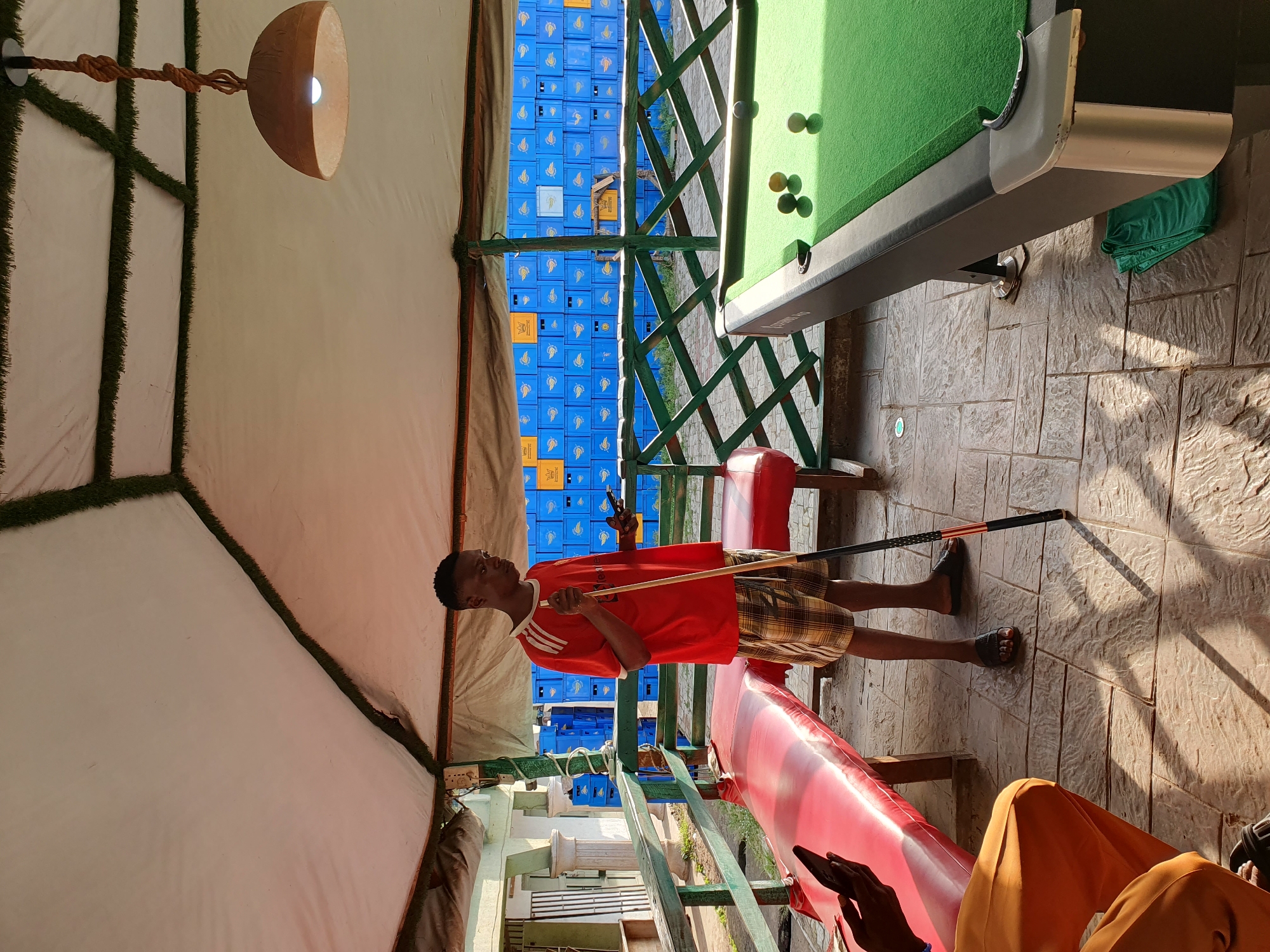 In Lokoja, Nigeria, a young man wearing a red t-shirt and checkered shorts stands near a pool table, holding a cue stick and a smartphone. He is positioned under a canopy with a white fabric roof adorned with artificial grass trim, supported by wooden beams. A pendant lamp made of a gourd hangs from the ceiling. In the background, rows of blue crates are stacked high, with some yellow crates interspersed. A red bench is visible to the left of the man, and to the right, the green surface of a pool table with several balls. The ground is paved with textured tiles, and shadows from the canopy and surrounding structures are cast across it. Another person, partially visible in orange clothing, sits on the left, holding a smartphone.