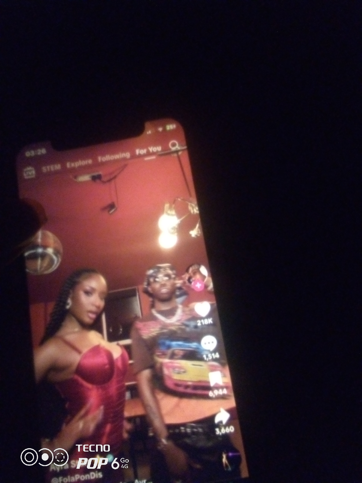 The image shows a close-up of a smartphone screen displaying a TikTok-like video interface. The video itself features two individuals, a woman on the left wearing a red corseted top and a man on the right wearing a patterned shirt and a headscarf. They appear to be posing. The background of the video is a red wall with some lighting fixtures and what looks like a disco ball on the left side.
On the smartphone screen's interface, text elements are visible, including "03:26", "STEM", "Explore", "Following", and "For You". There are also icons for likes, comments, and shares, with accompanying numbers like "218K", "1,514", "6,944", and "3,660". At the bottom of the screen, there is a logo that reads "TECNO POP 6 4G" and a username "@FolaPonDis".
The image is captured in a dark environment, with the phone screen being the primary light source. There is no discernible information in the image that can be used to determine the specific location within the city of Howayi, Nigeria. The content displayed is digital and does not provide geographic context.