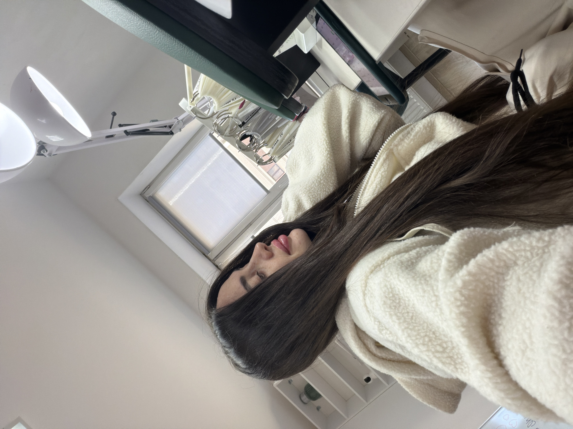 A woman with long brown hair, wearing a white sherpa jacket, is lying on a bed or treatment table. Her face is turned to the side, partially obscured by her hair. Above her is an adjustable white lamp with two lights. A window is visible in the background, as well as a display of nail polish swatches hanging above her. The location cannot be determined as being in Saint Petersburg, Russia from the visual information.