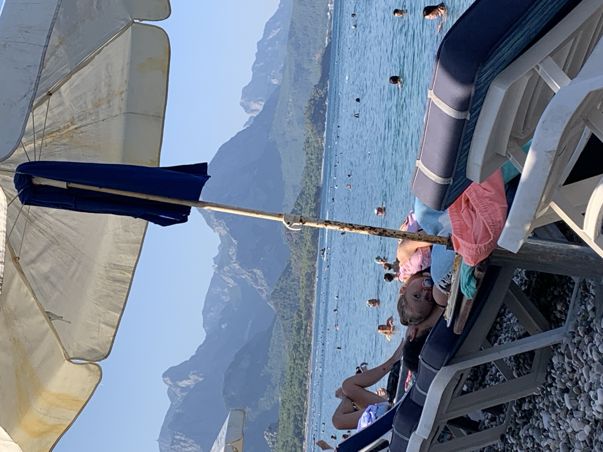 The image shows a crowded beach in Kemer, Turkey, with numerous people swimming and sunbathing. In the foreground, several beach chairs are set up on a pebble beach, with some occupied by individuals relaxing under a large umbrella. A child with a pacifier is lying on a chair in the center foreground, with another person next to them. The sea is calm and blue, with many people in the water. In the background, lush green hills and majestic mountains form a scenic backdrop to the beach. The sky is clear and blue, indicating a sunny day.