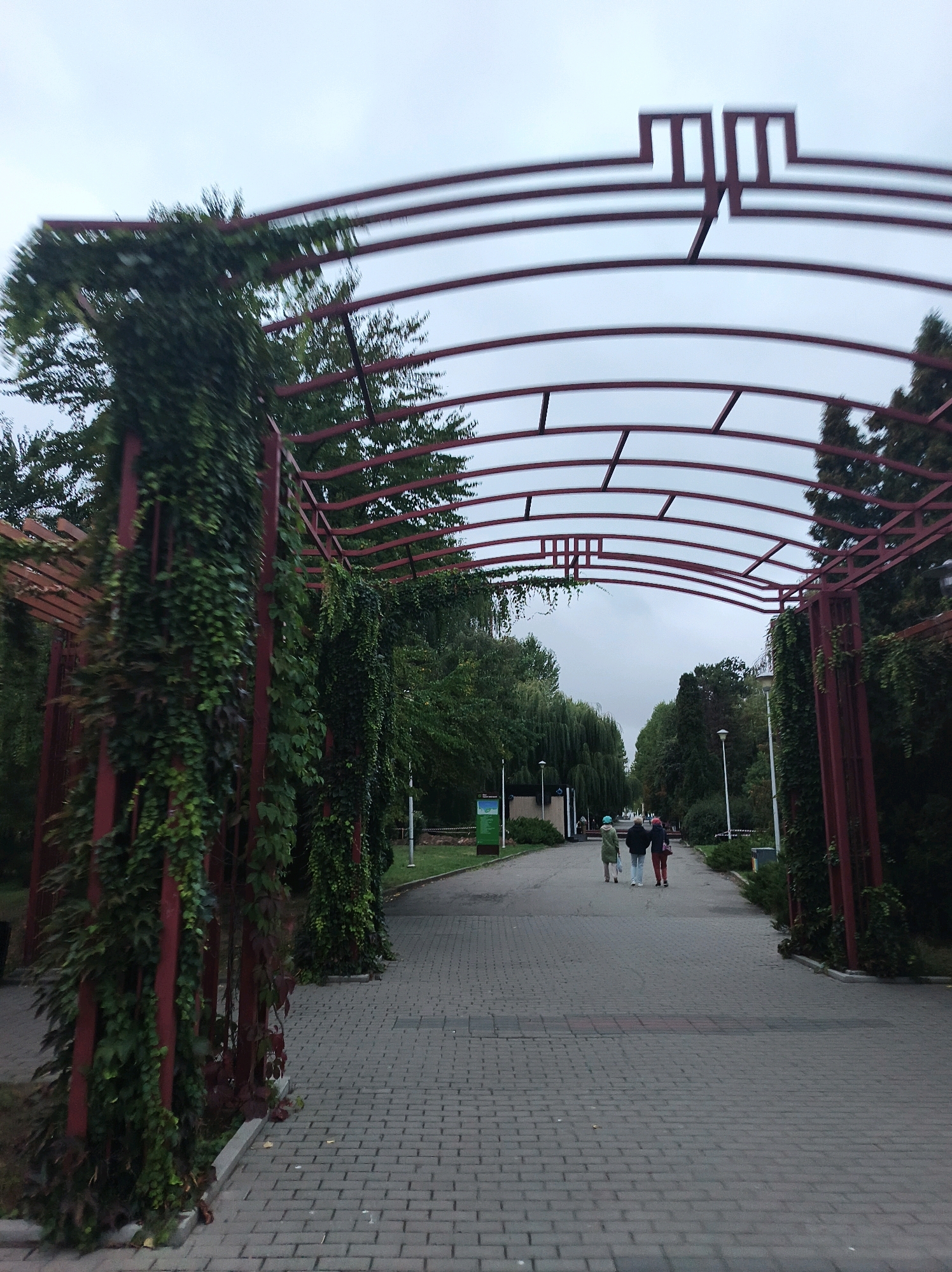 The image shows a pathway in a park in Ternopil, Ukraine. The pathway is paved with grey bricks and lined with green bushes and trees. An arched metal structure, painted red and covered with ivy, spans the pathway. Three people are walking away from the camera on the pathway. The sky is overcast.
