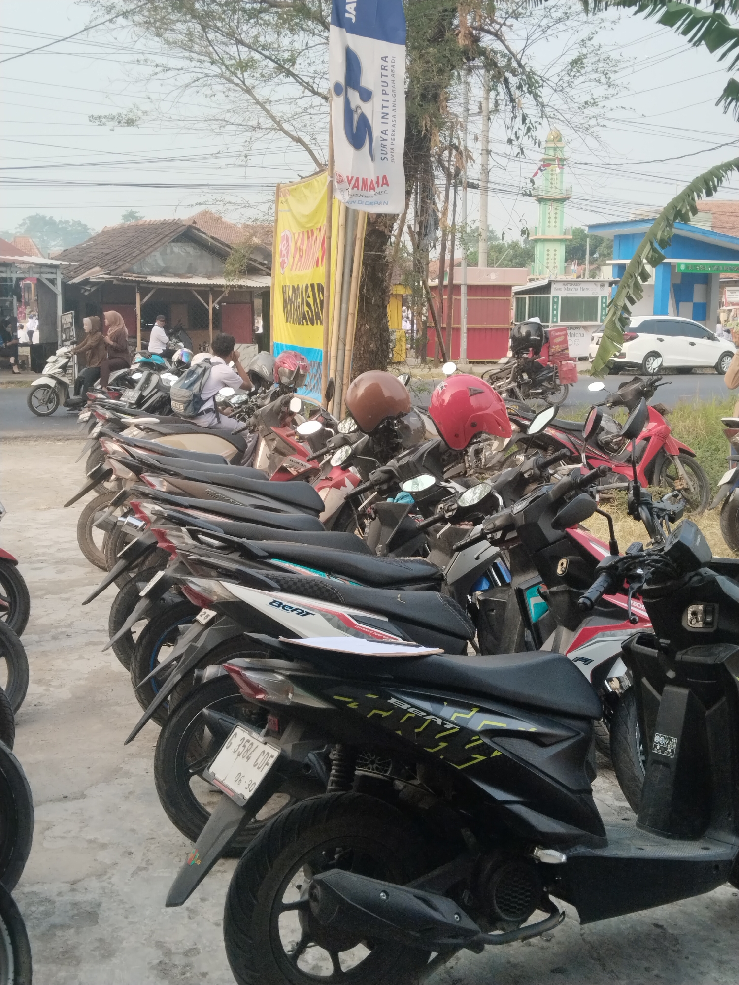 The image depicts a street scene in Pakulaut, Indonesia, with a prominent row of parked motorcycles in the foreground. A white car is visible in the background, along with a green minaret of a mosque. Several buildings line the street, including what appears to be a small shop or eatery. A banner with text in Indonesian, likely advertising a product or event, hangs from a pole. Two women wearing headscarves are seated on a parked motorcycle. The overall atmosphere suggests a typical day in a bustling Indonesian town. The scene is captured from a low angle, emphasizing the motorcycles. The lighting suggests it is daytime, likely late afternoon due to the soft, warm light.