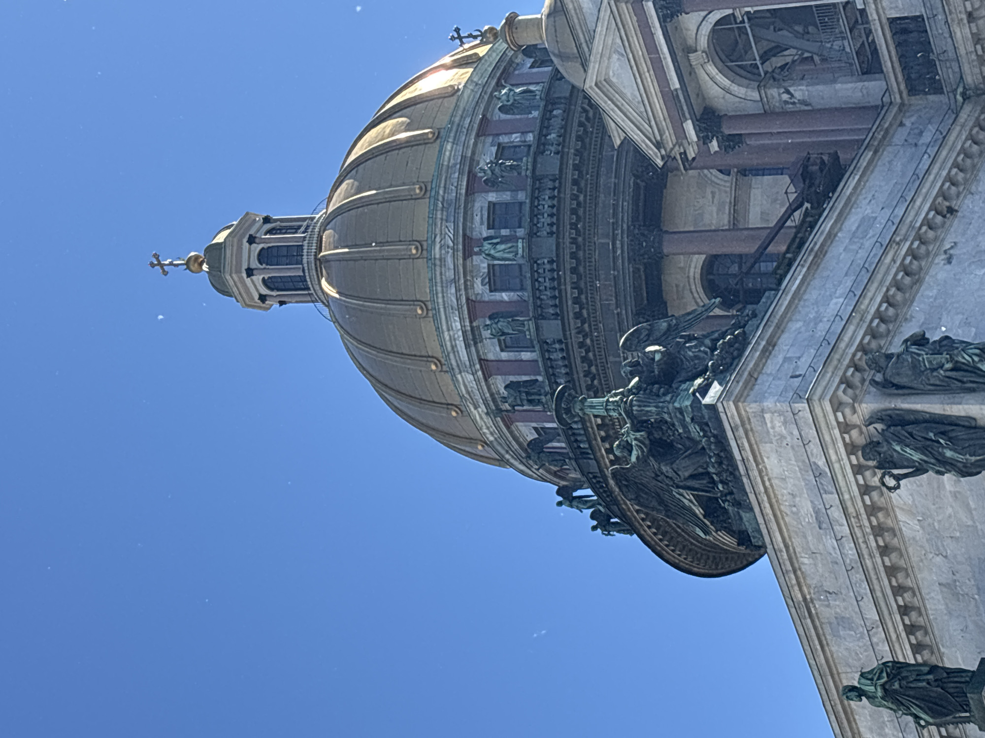 This is a close-up, low-angle shot of the dome and upper structure of Saint Isaac's Cathedral in Saint Petersburg, Russia, against a clear blue sky. The prominent golden dome is segmented and topped with a lantern featuring arched windows and a cross. Sculptural elements, including what appear to be angels or figures with wings, are visible on the exterior ledges and corners of the building. Additional statues of robed figures are positioned on the lower marble facade. The architecture exhibits classical influences with columns and ornate detailing. The overall impression is one of grandeur and historical significance.